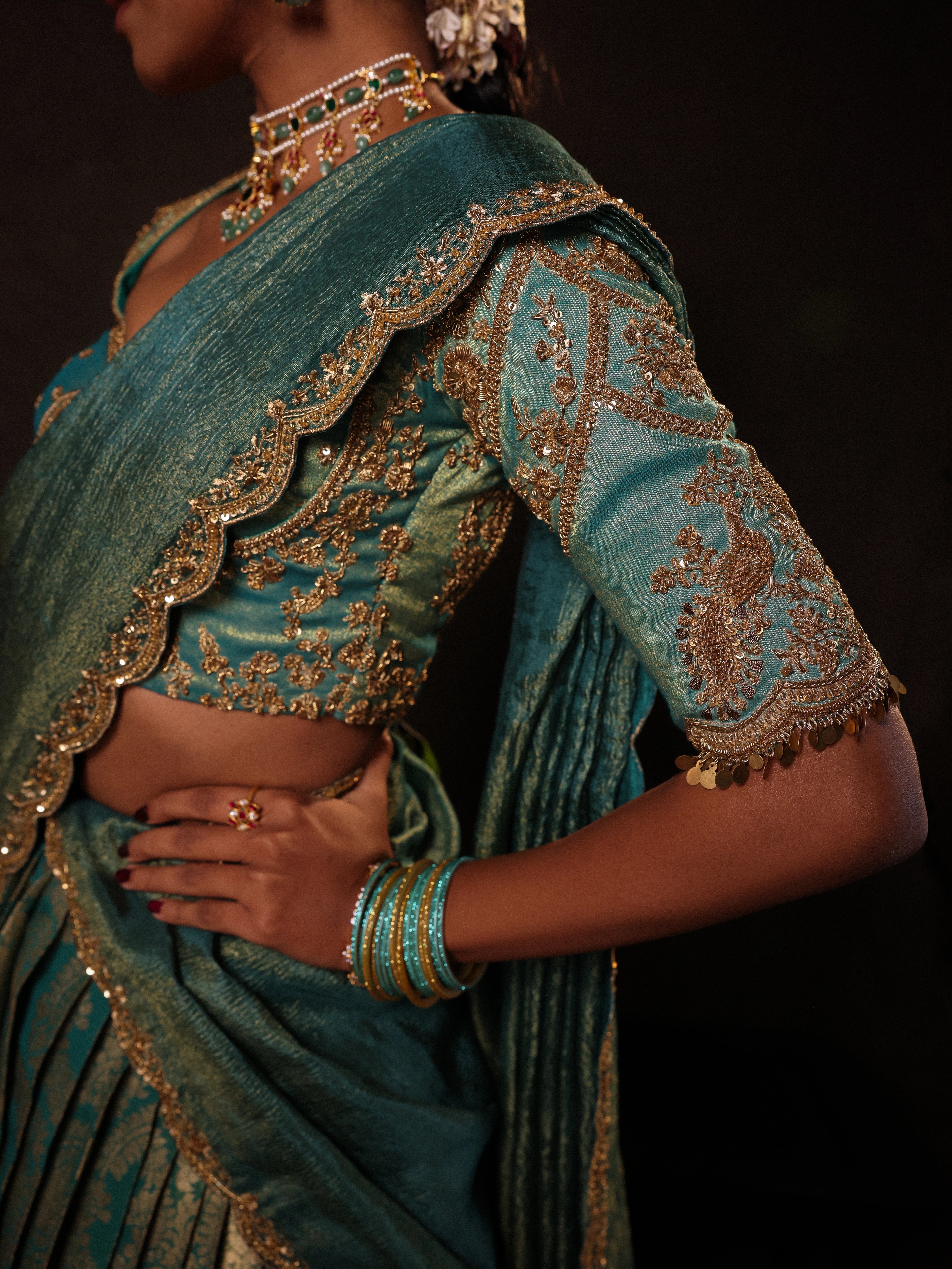 Model Gayathri Dhanasekar showcasing detailing on blouse in a teal half saree (silk blouse and skirt and tissue dupatta) handcrafted with zardozi, zari, cutdana, sequins and metal coin hangings. Team Archana Karthick completed the style with teal glass bangles, a ring, a choker, jhumkas, nose ring and a maang tikka.