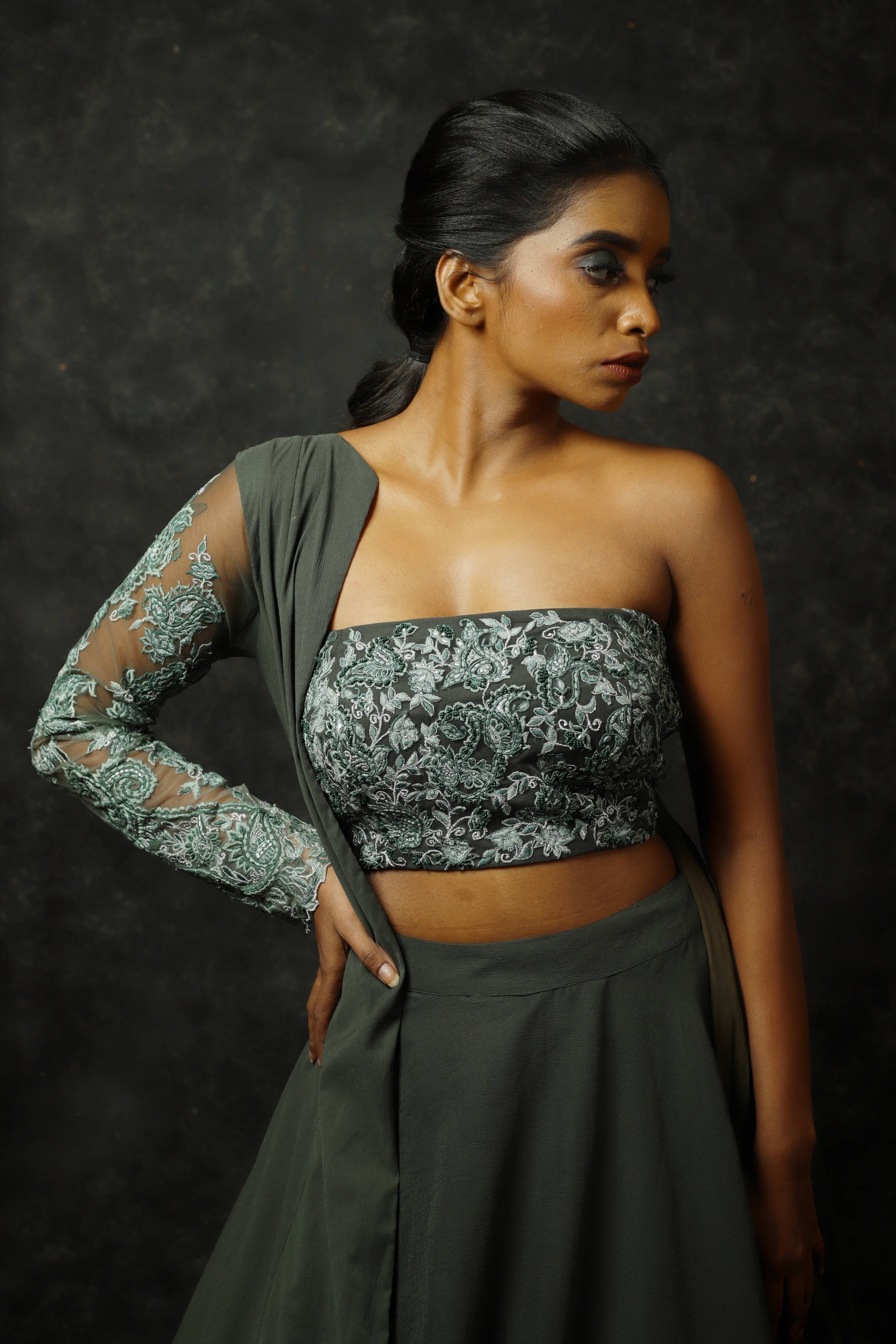 Model Posing facing front but looking to her left in an olive green georgette masakali lehenga with long shrug and bralette. She is making sure the work on the bralette and the sleeve is clearly seen.