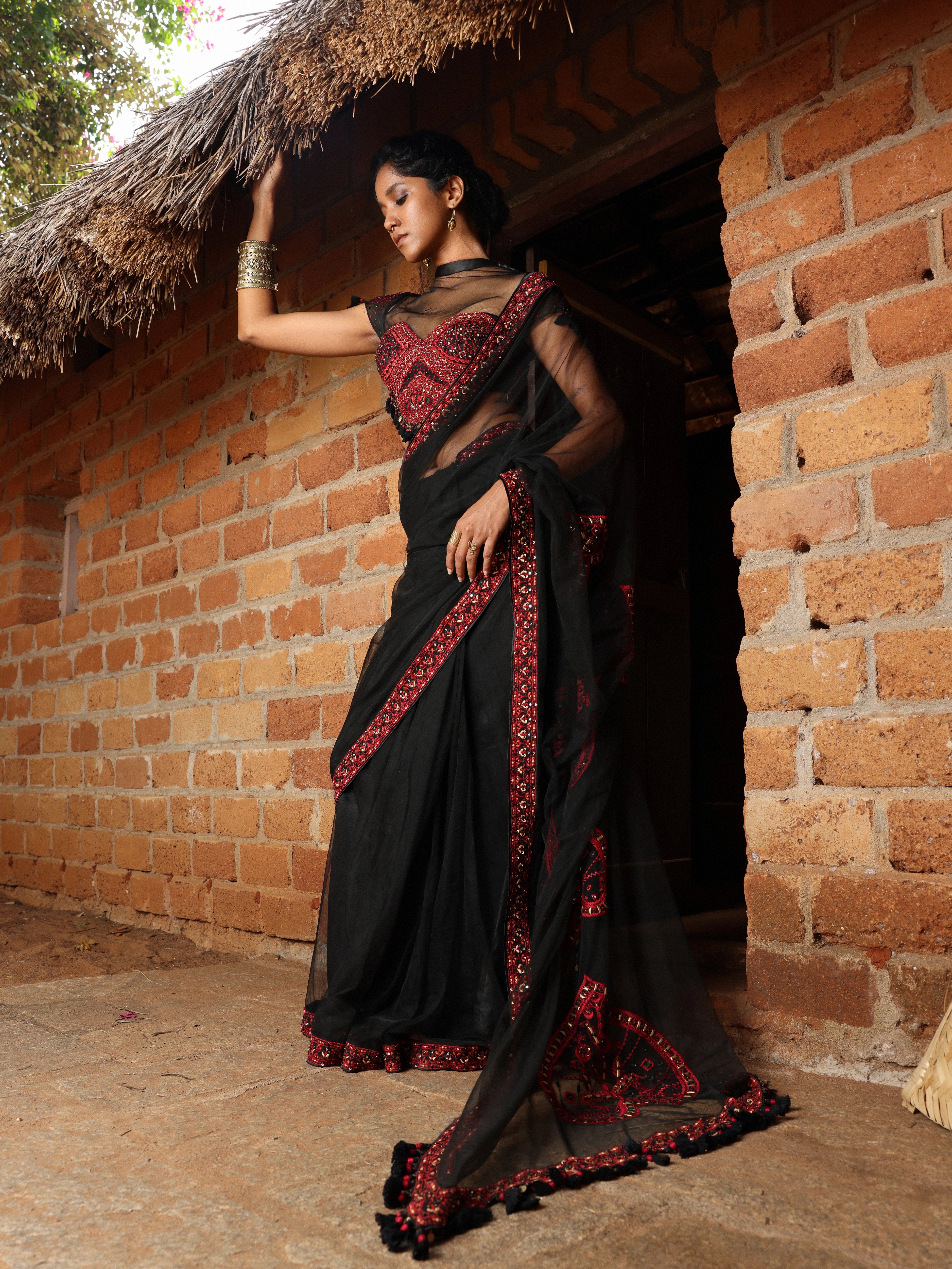 Model in black and red net saree with blouse. Posing outside a hut.