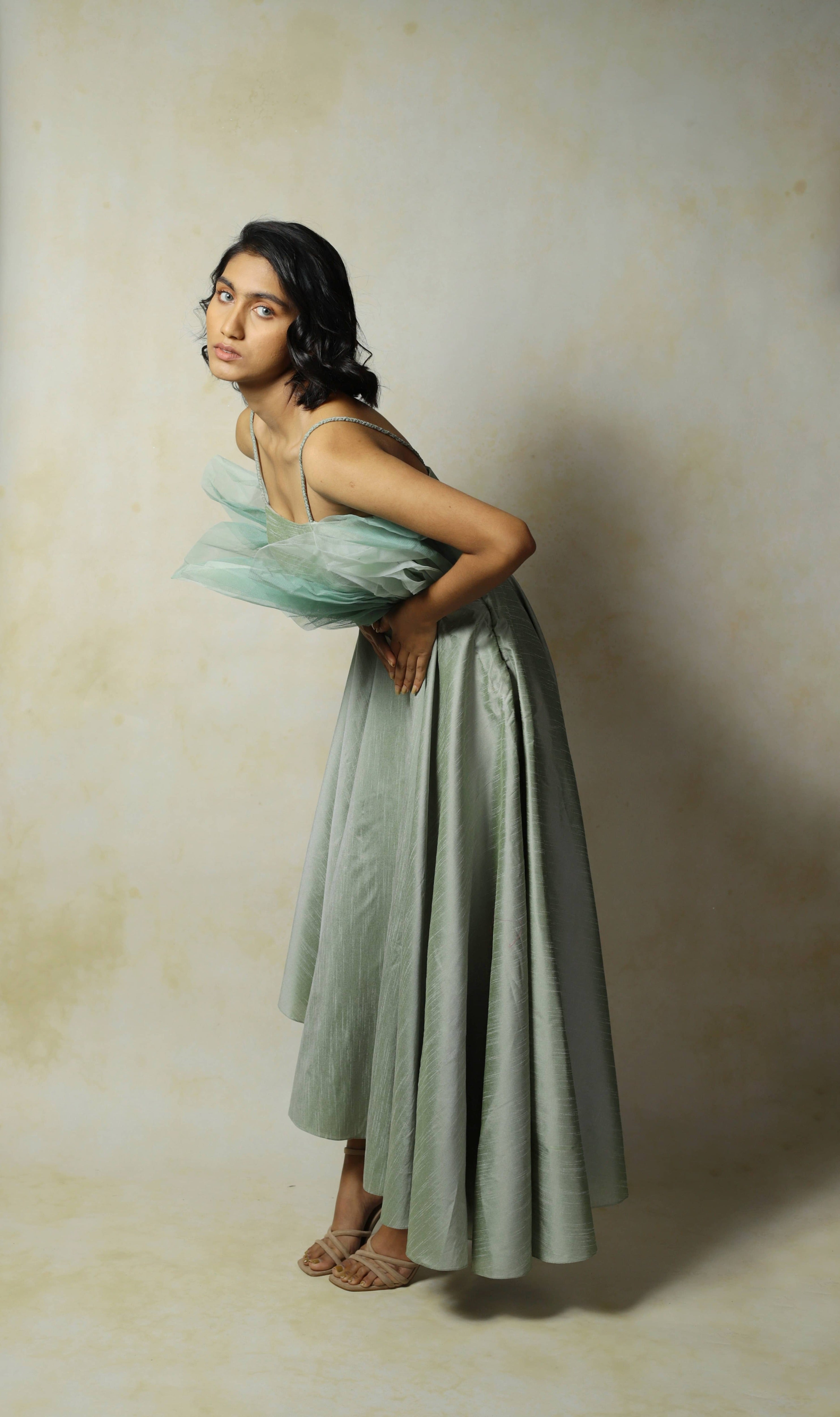 Model posing bent forward and looking to her left in a green raw silk and structured net dress designed by Archana Karthick.