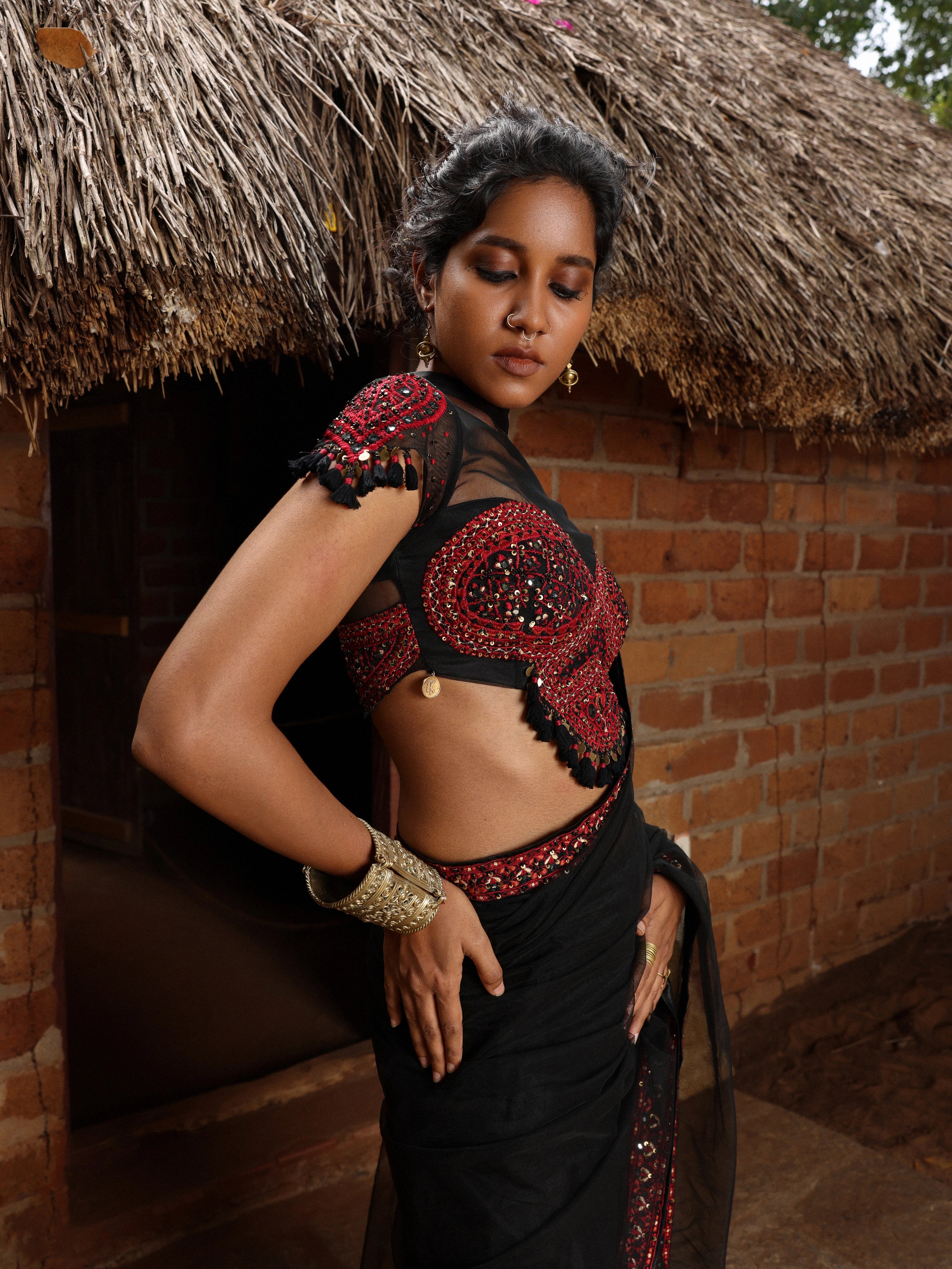 Model posing in black and red net saree with blouse. Outside a hut.