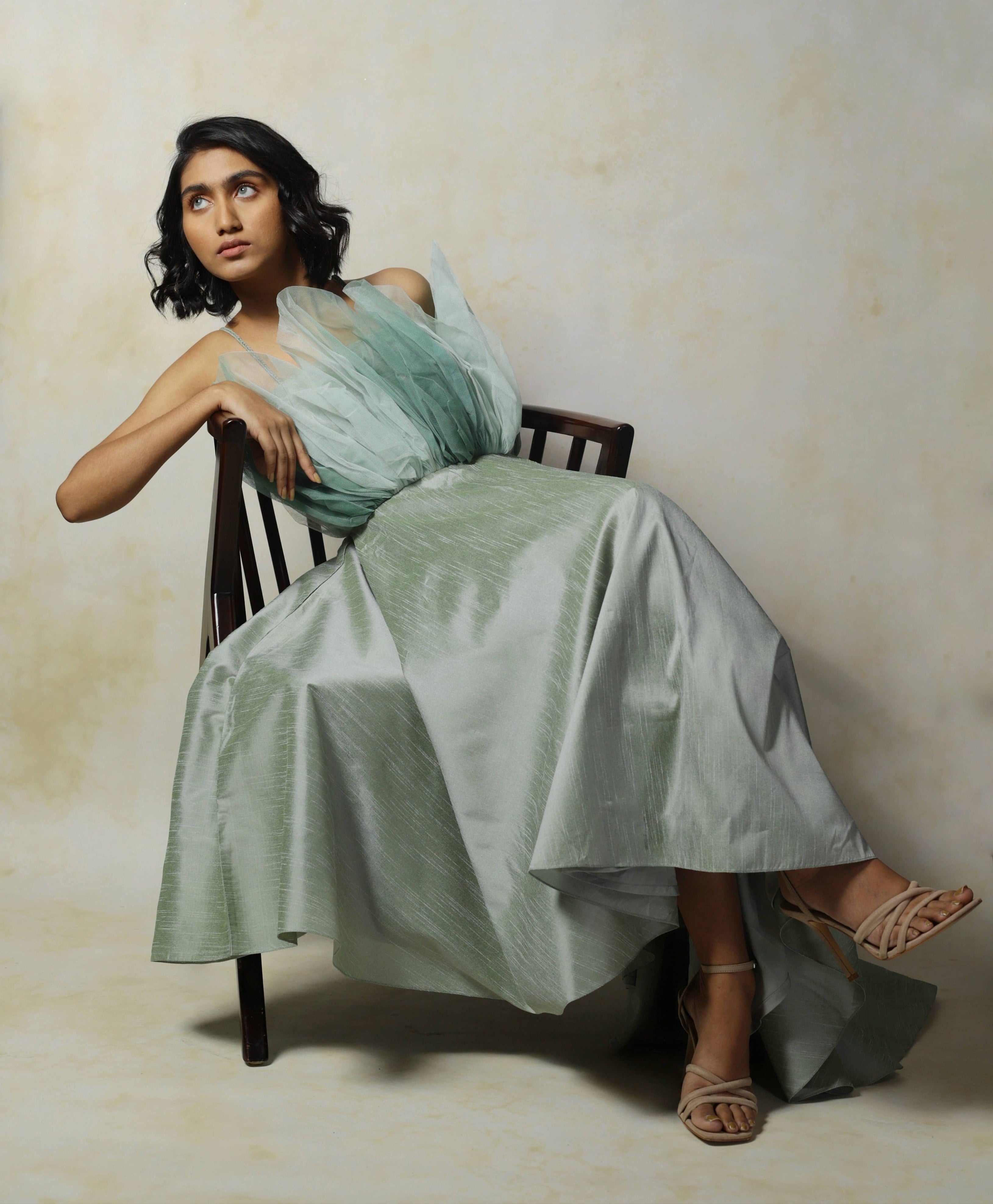 Model posing seated in a corporate stylr and looking to her right in a green raw silk and structured net dress designed by Archana Karthick.