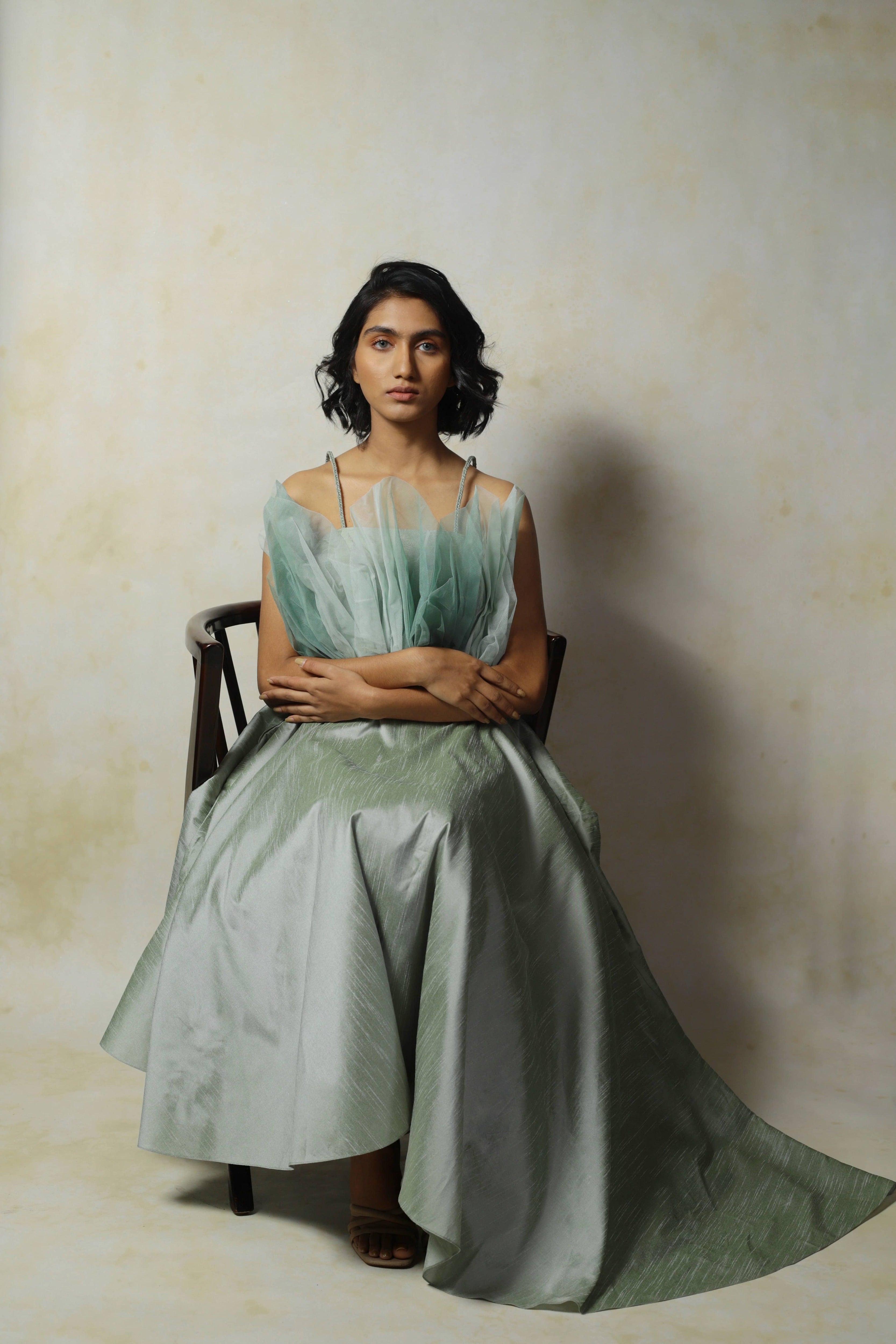 Model posing seated straight backed and looking dead ahead in a green raw silk and structured net dress designed by Archana Karthick.