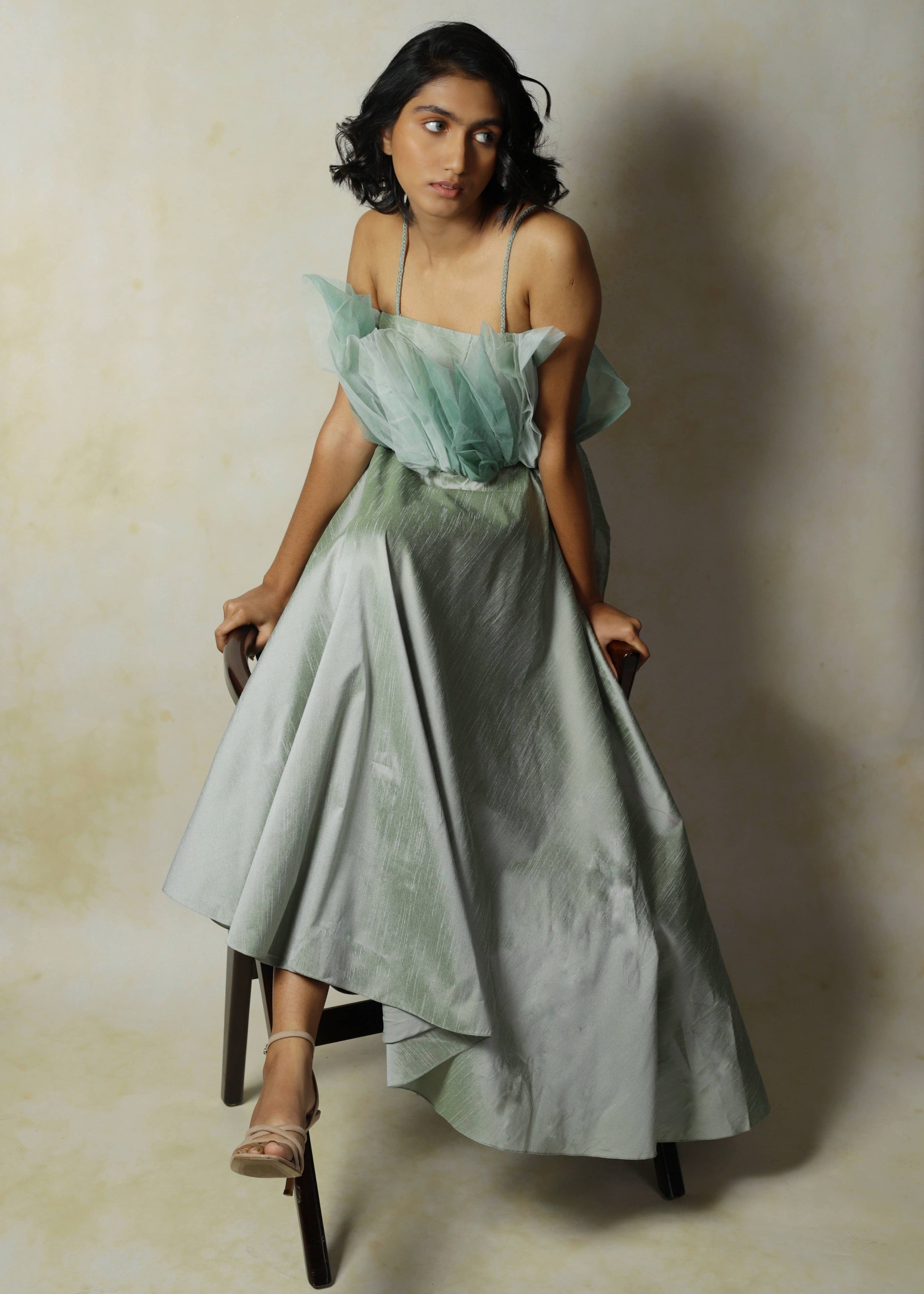 Model posing stylishly seated obliquely and looking to her left in a green raw silk and structured net dress designed by Archana Karthick.
