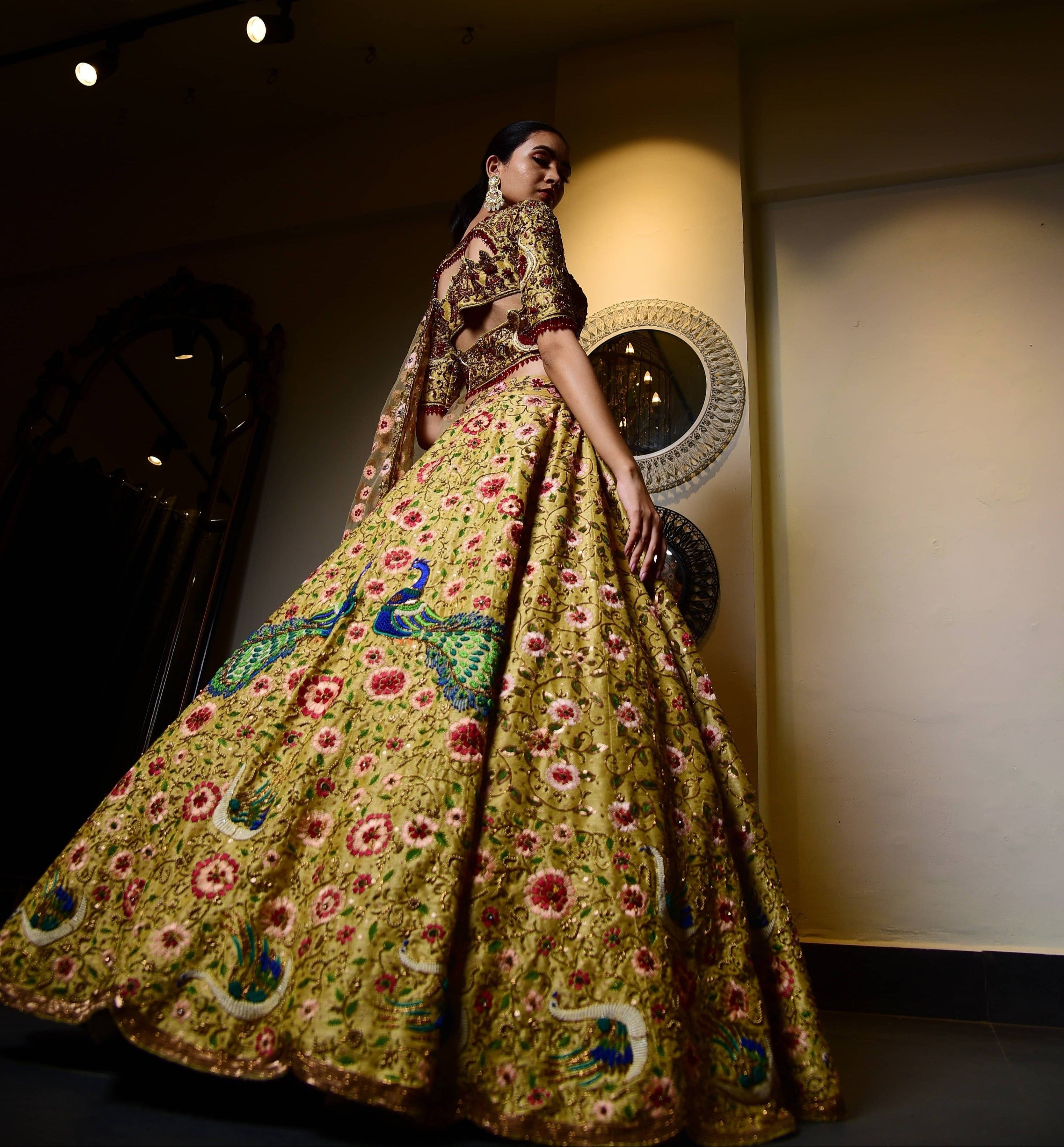 The model is posing by looking back giving us the full rear view of the outfit. The raw silk lehenga with heavy intricate resham, sequins, beads, stonework embellishments all over the lehenga, palanquin concept blouse and tulle dupatta.