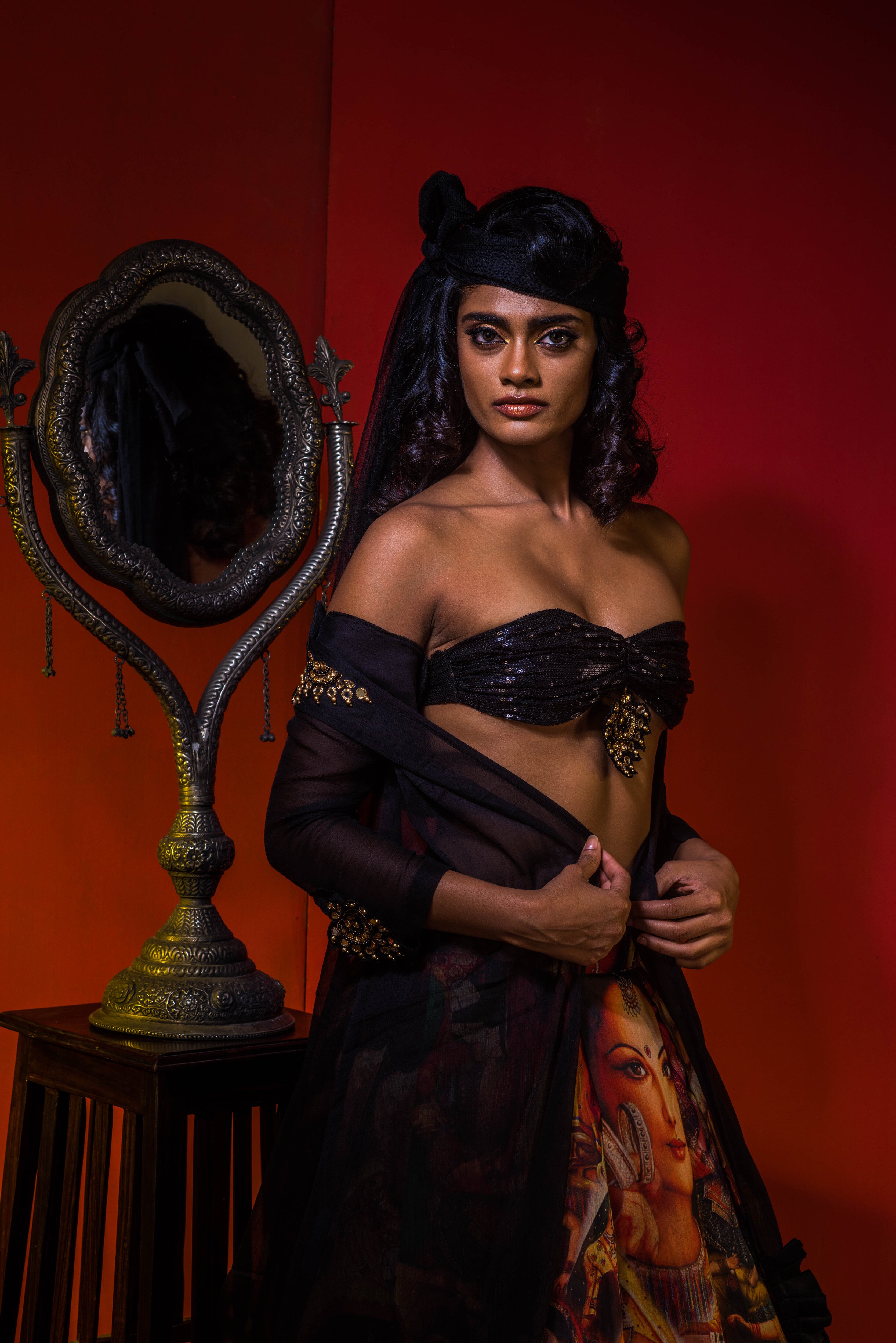 Supermodel Archana Akil Kumar in a heavily embellished black georgette bralette and long shrug paired with a printed colorful skirt looking staright into the lens with the shrug pulled down to her waist revealing the bralette and looking sideways.