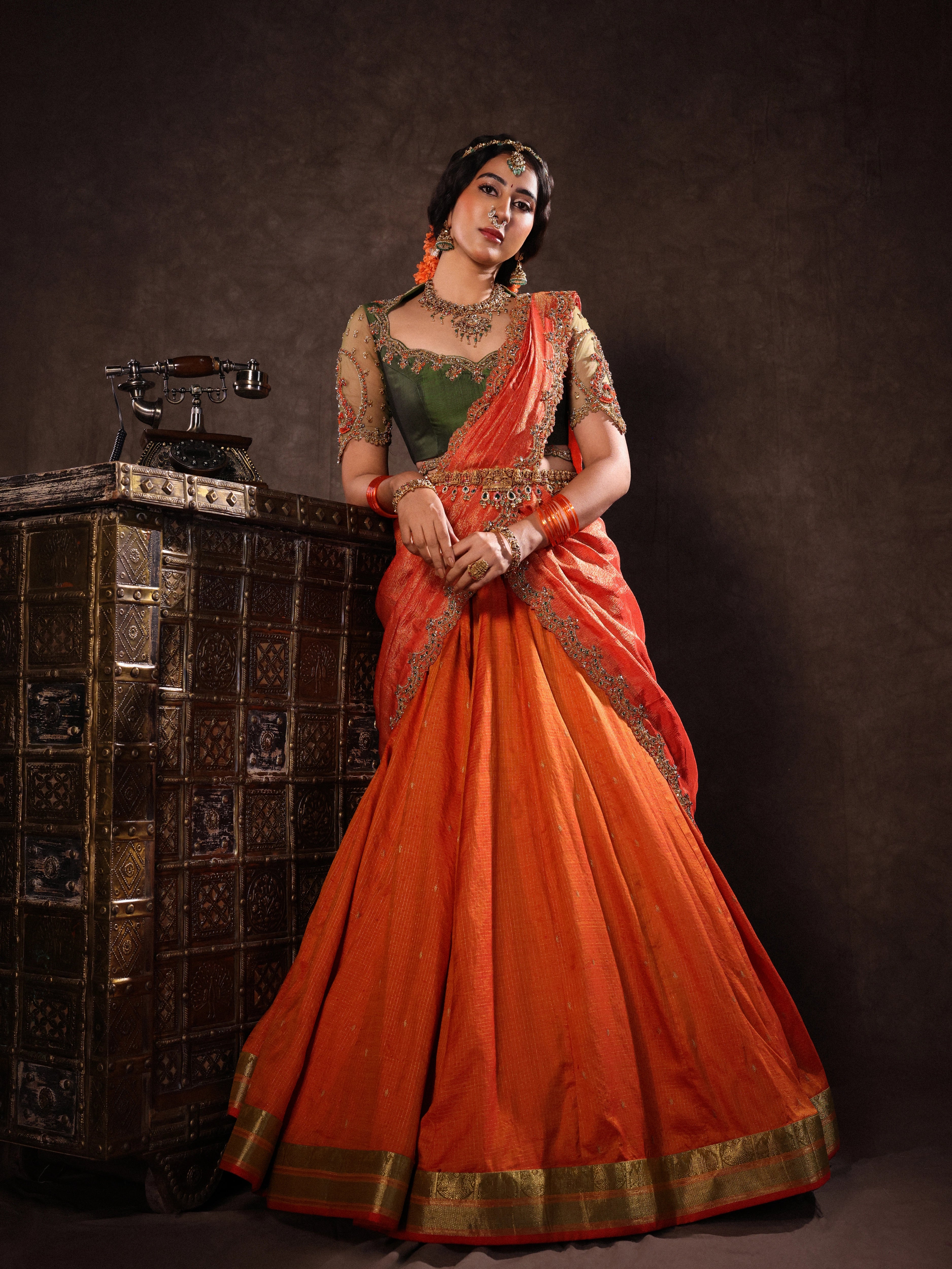 Actress Akshaya Hariharan in an orange & olive green silk half saree set with tulle sleeves. Handcrafted with zardozi, resham, cutdana, sequins & rhinestones. Team Archana Karthick completed the style with orange glass bangles, a ring, jhumkas, nose ring and a maang tikka.