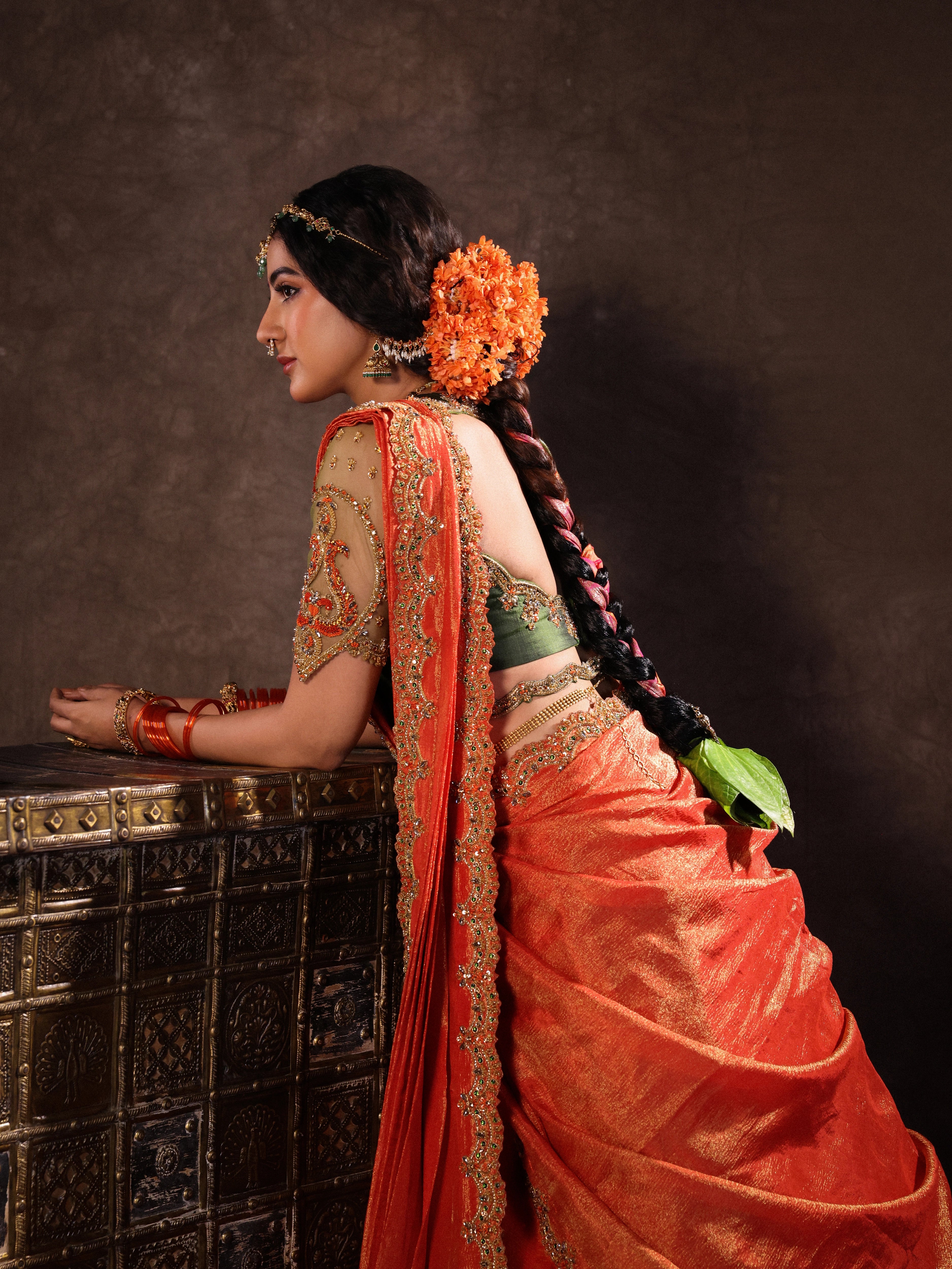 Actress Akshaya Hariharan leaning in an orange & olive green silk half saree set with tulle sleeves. Handcrafted with zardozi, resham, cutdana, sequins & rhinestones. Team Archana Karthick completed the style with orange glass bangles, a ring, jhumkas, nose ring and a maang tikka.