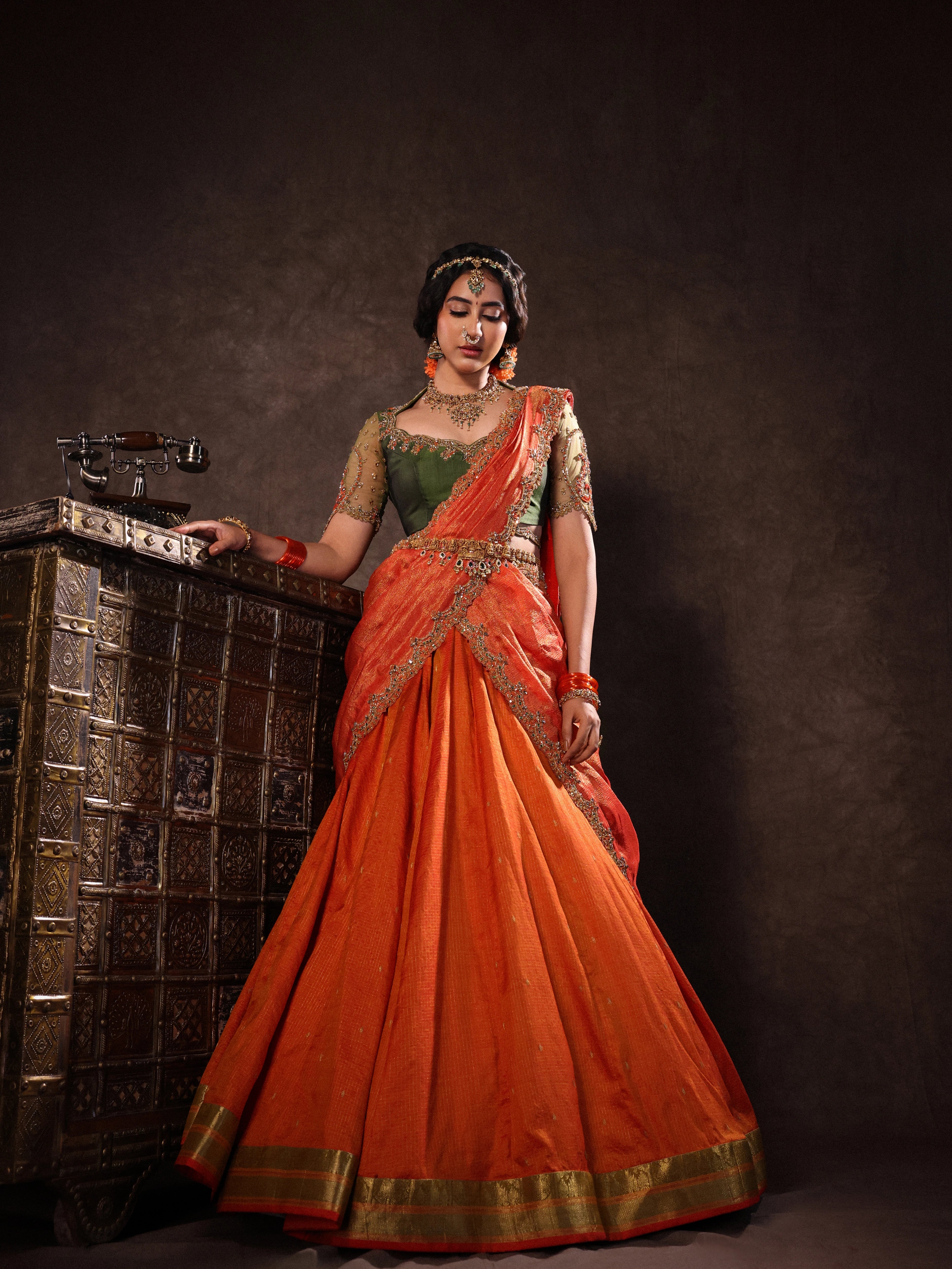 Actress Akshaya Hariharan looking down in an orange & olive green silk half saree set with tulle sleeves. Handcrafted with zardozi, resham, cutdana, sequins & rhinestones. Team Archana Karthick completed the style with orange glass bangles, a ring, jhumkas, nose ring and a maang tikka.