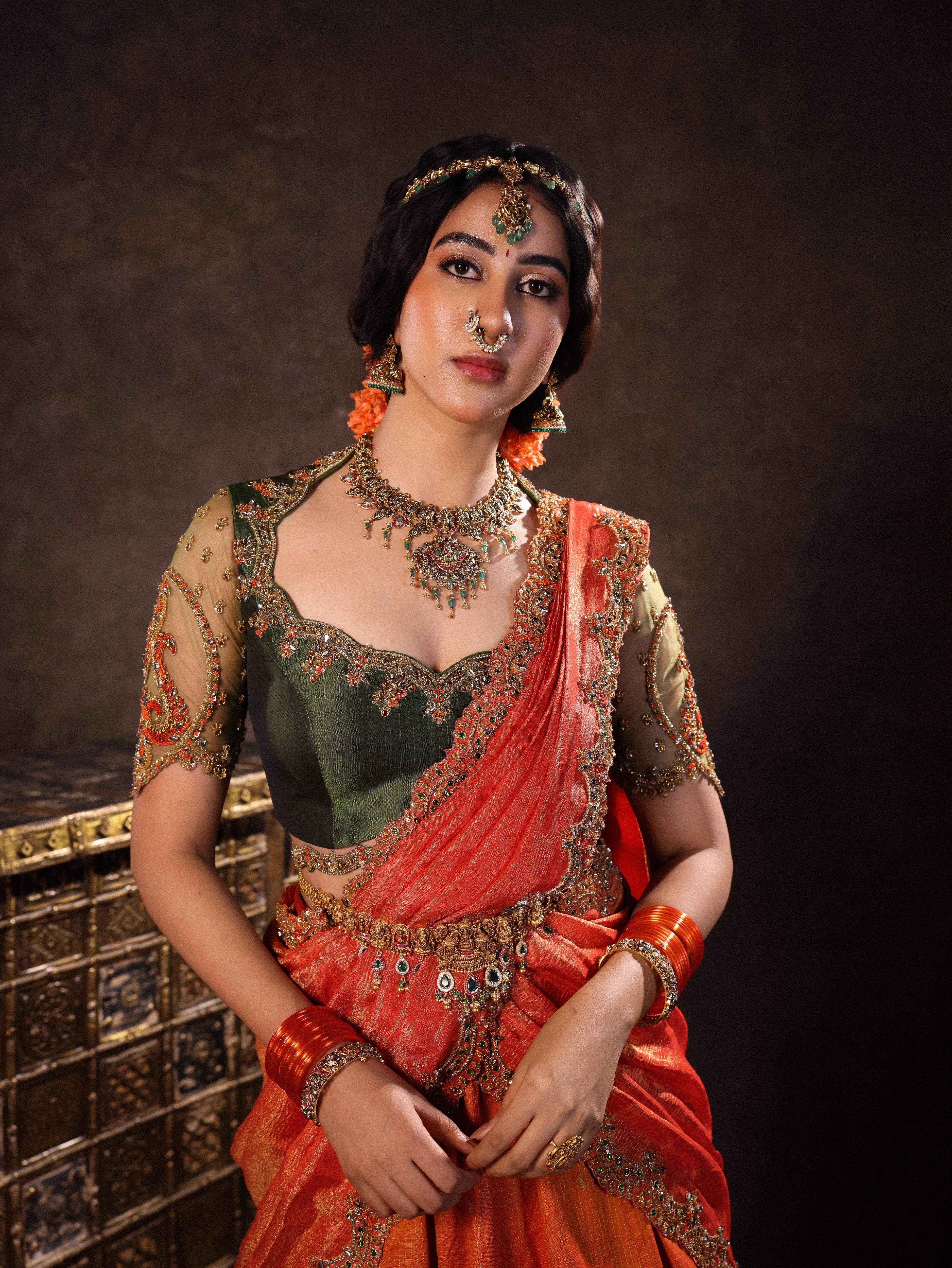 Actress Akshaya Hariharan poses in an orange & olive green silk half saree set with tulle sleeves. Handcrafted with zardozi, resham, cutdana, sequins & rhinestones. Team Archana Karthick completed the style with orange glass bangles, a ring, jhumkas, nose ring and a maang tikka.