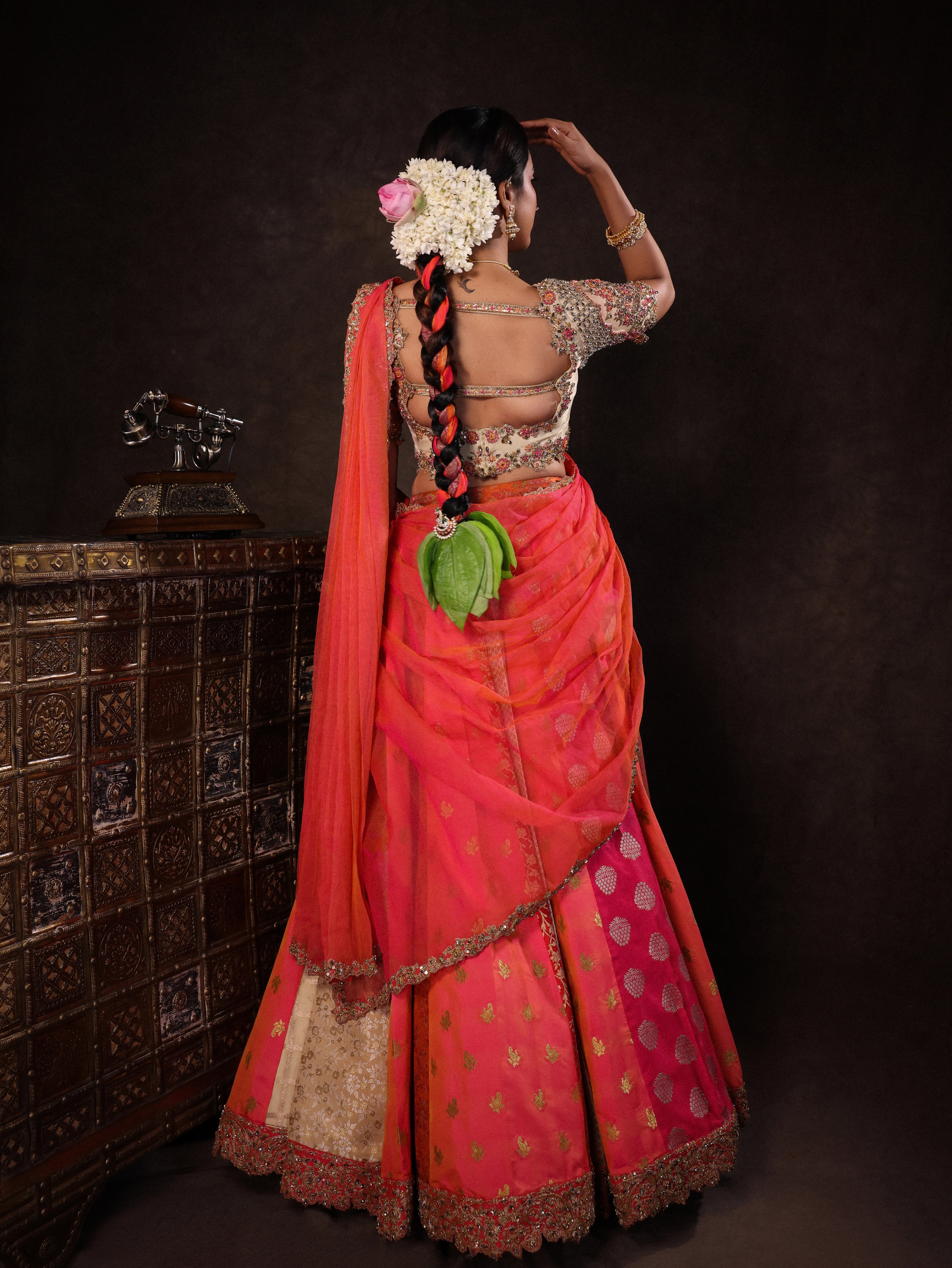 Actress Akshaya Hariharan posing for back detailing in an ivory & pink brocade half saree set with tulle sleeves & dupatta. Team Archana Karthick completed the style with purple glass bangles, a ring, jhumkas, nose ring and a maang tikka.