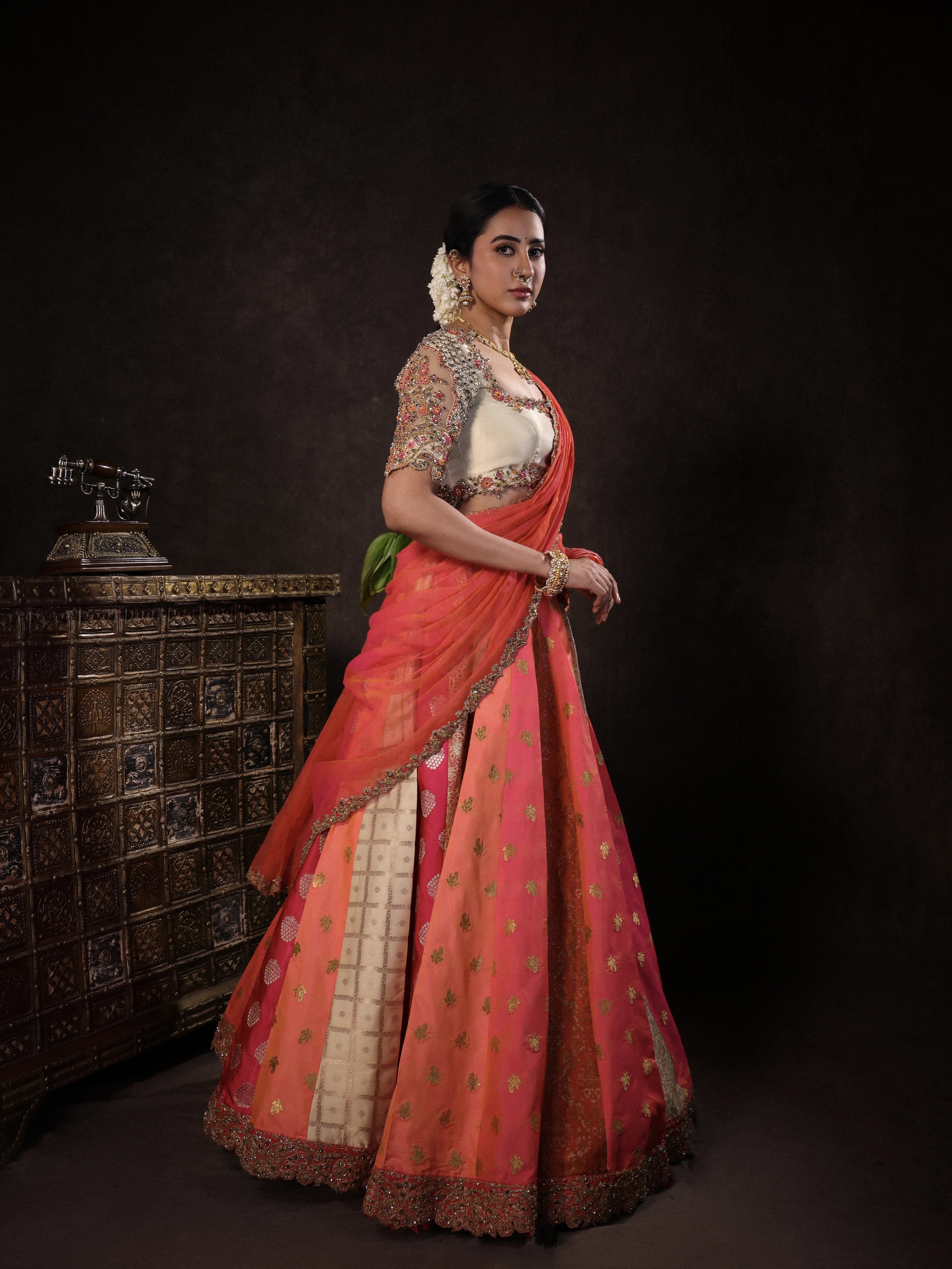 Actress Akshaya Hariharan posing for side profile in an ivory & pink brocade half saree set with tulle sleeves & dupatta. Team Archana Karthick completed the style with purple glass bangles, a ring, jhumkas, nose ring and a maang tikka.