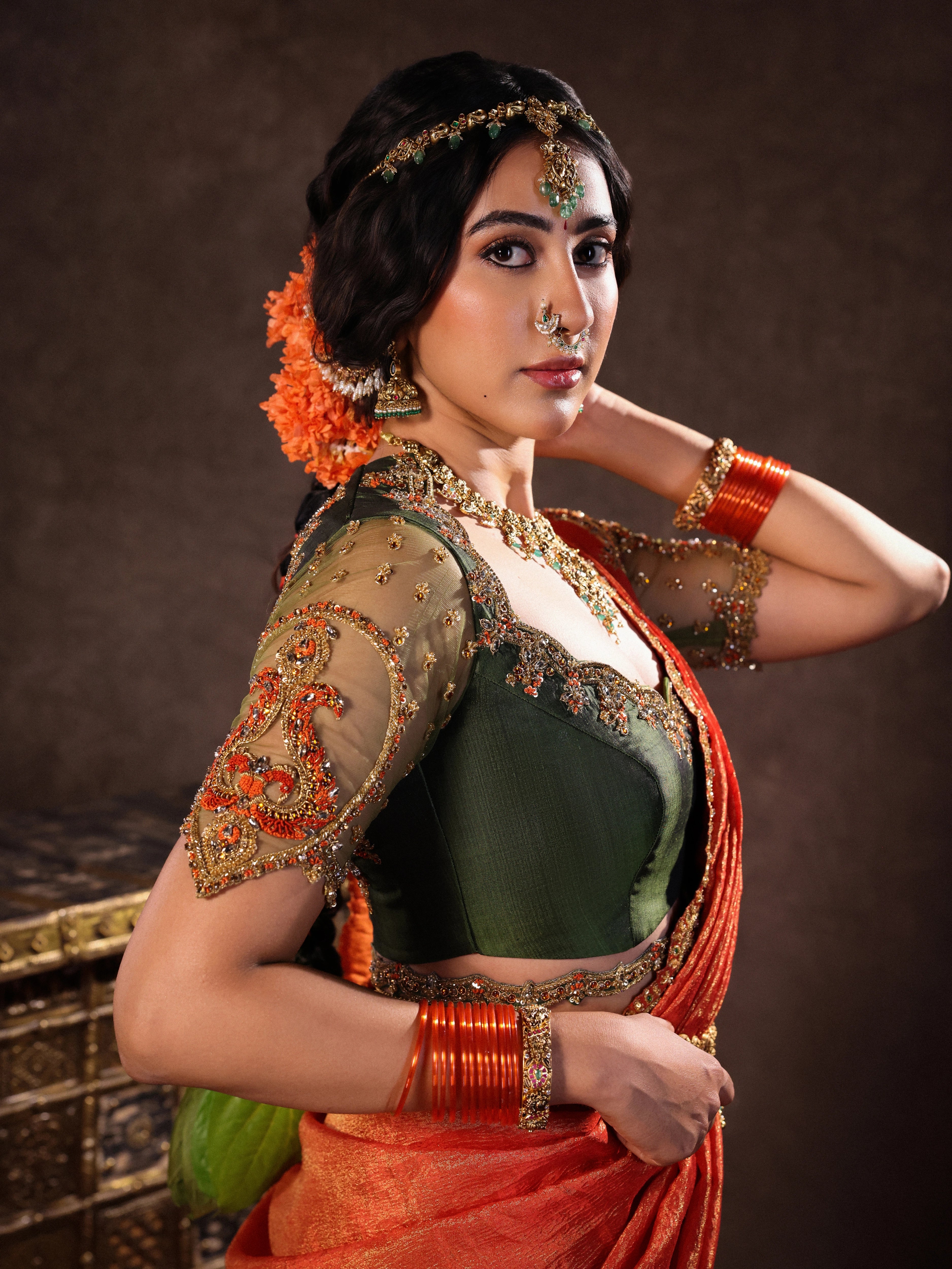 Actress Akshaya Hariharan posing in an orange & olive green silk half saree set with tulle sleeves. Handcrafted with zardozi, resham, cutdana, sequins & rhinestones. Team Archana Karthick completed the style with orange glass bangles, a ring, jhumkas, nose ring and a maang tikka.