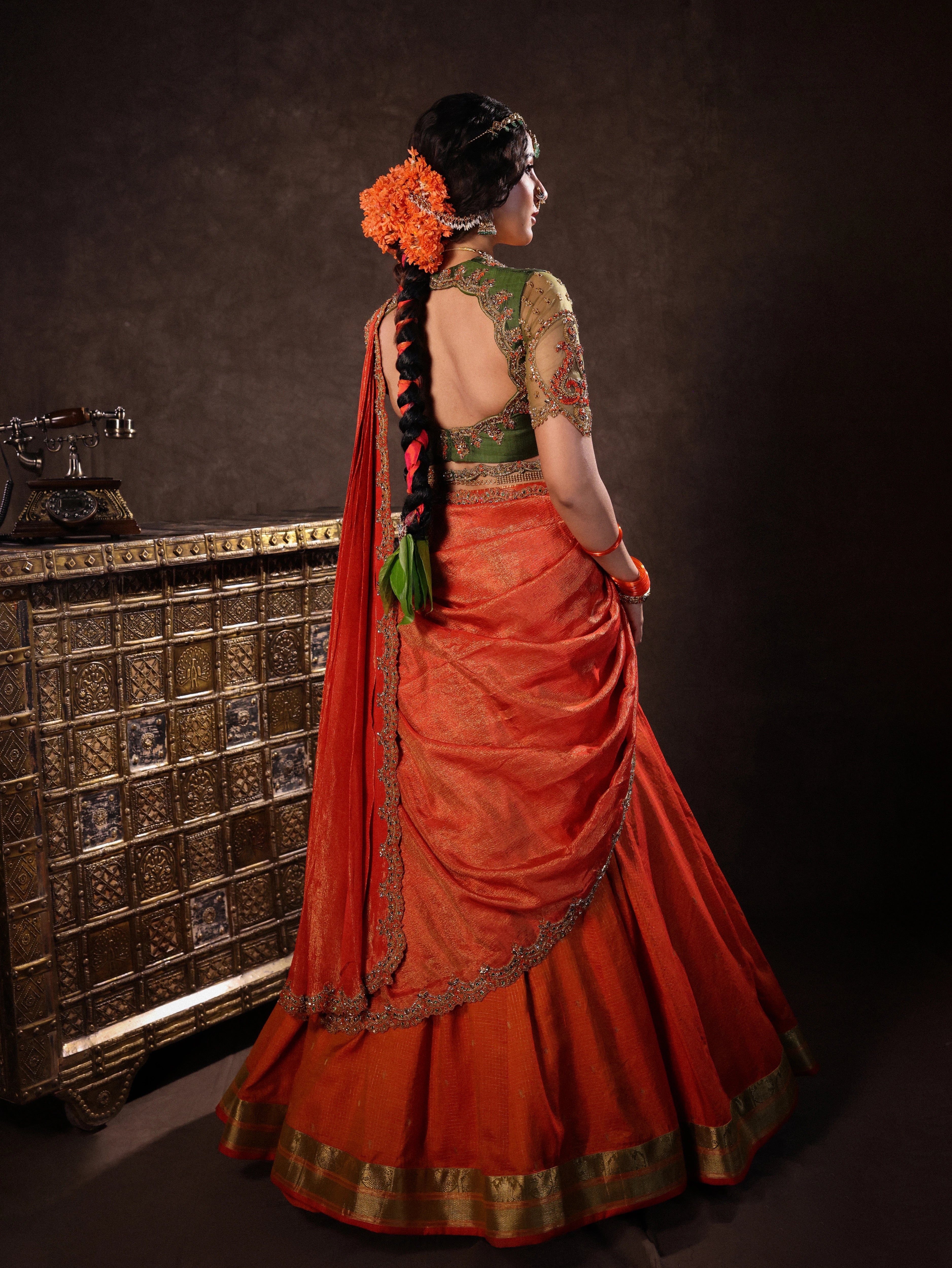 Actress Akshaya Hariharan showcasing back detaIling in an orange & olive green silk half saree set with tulle sleeves. Handcrafted with zardozi, resham, cutdana, sequins & rhinestones. Team Archana Karthick completed the style with orange glass bangles, a ring, jhumkas, nose ring and a maang tikka.