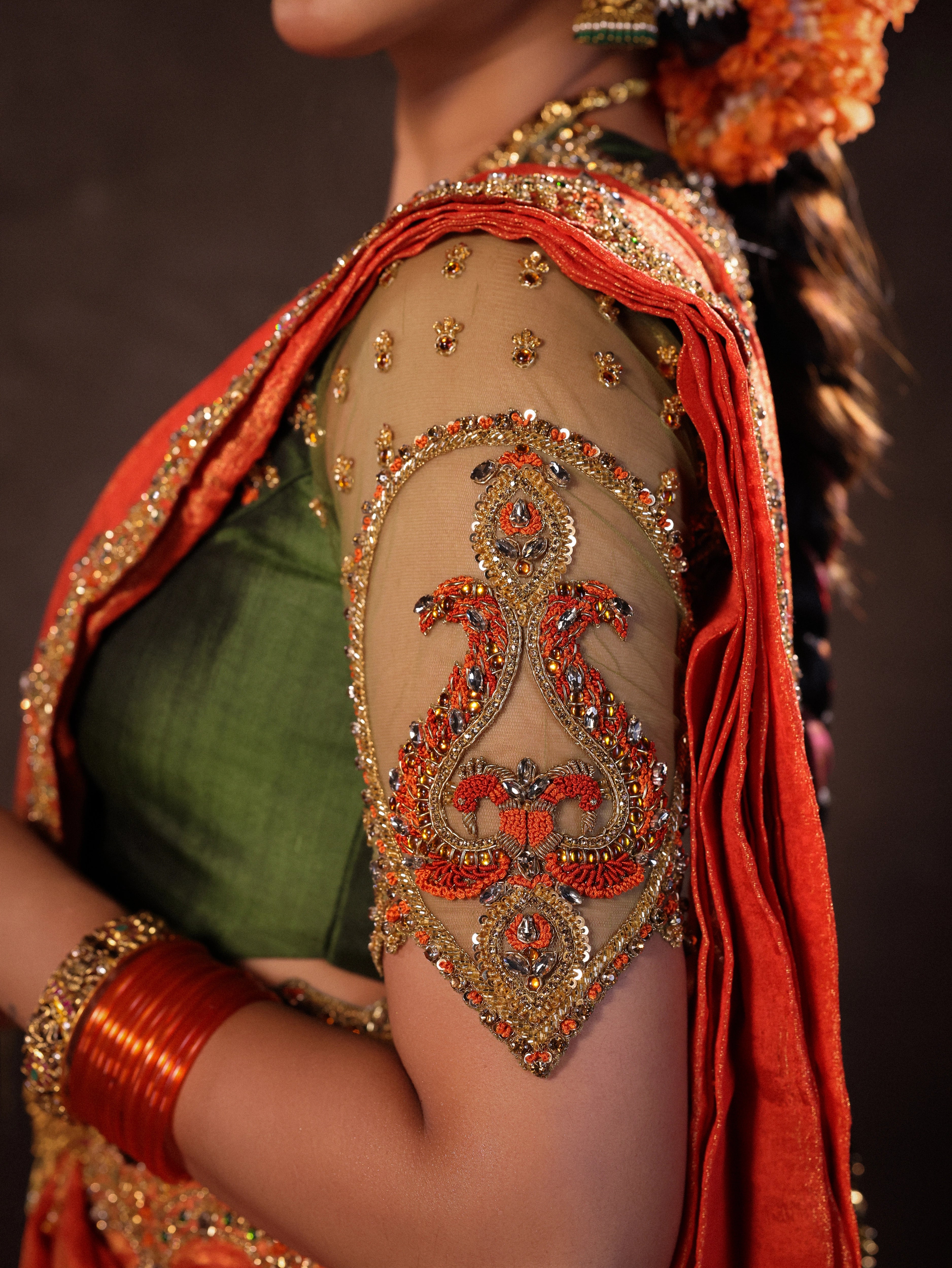 Actress Akshaya Hariharan showcasing sleeve detaIling in an orange & olive green silk half saree set with tulle sleeves. Handcrafted with zardozi, resham, cutdana, sequins & rhinestones. Team Archana Karthick completed the style with orange glass bangles, a ring, jhumkas, nose ring and a maang tikka.