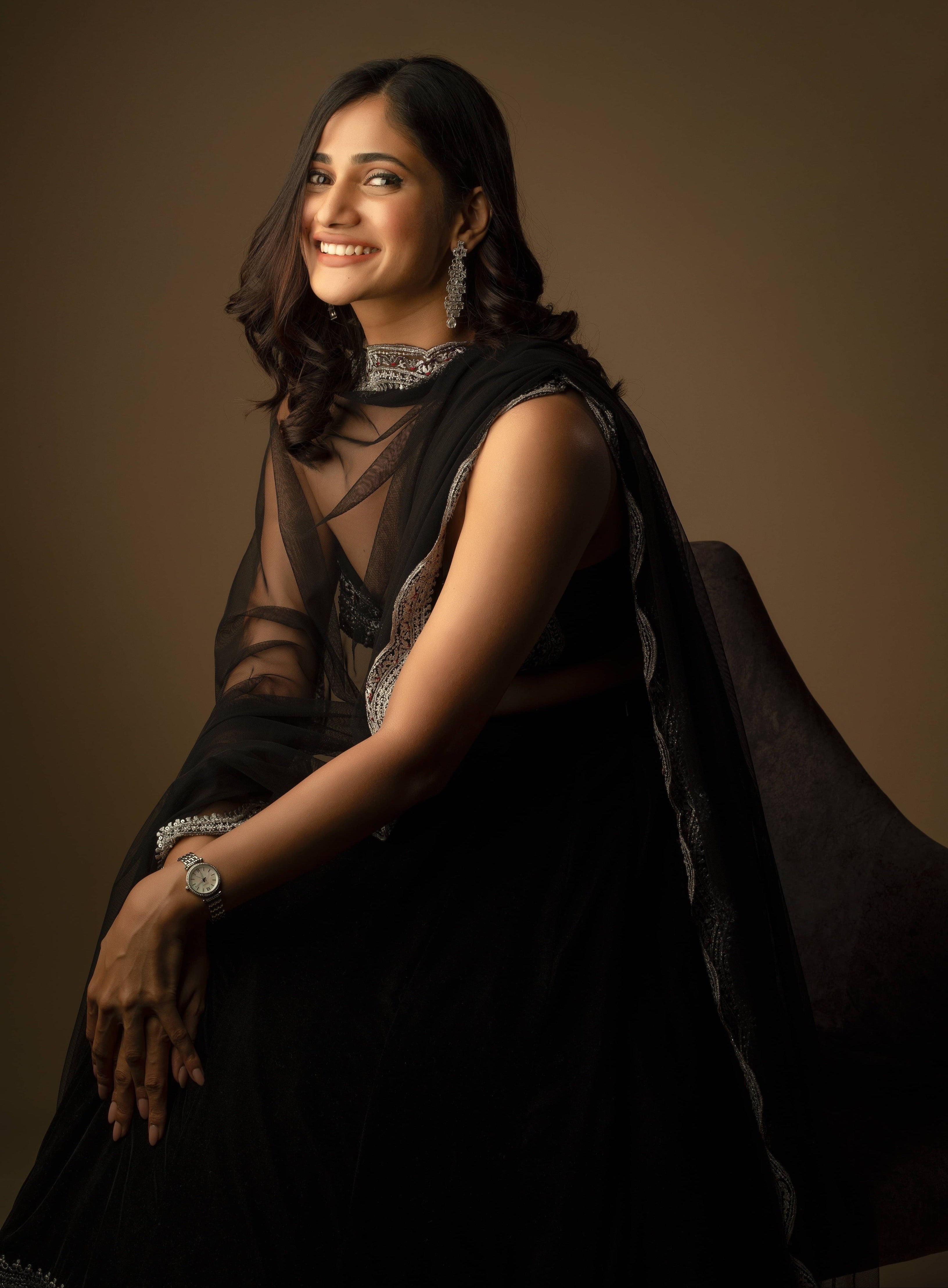 Bigg Boss fame and actress Losliya Mariyanesan in black velvet lehenga with silver embellishments on the borders and bralette. Posing seated and smiling at the camera.