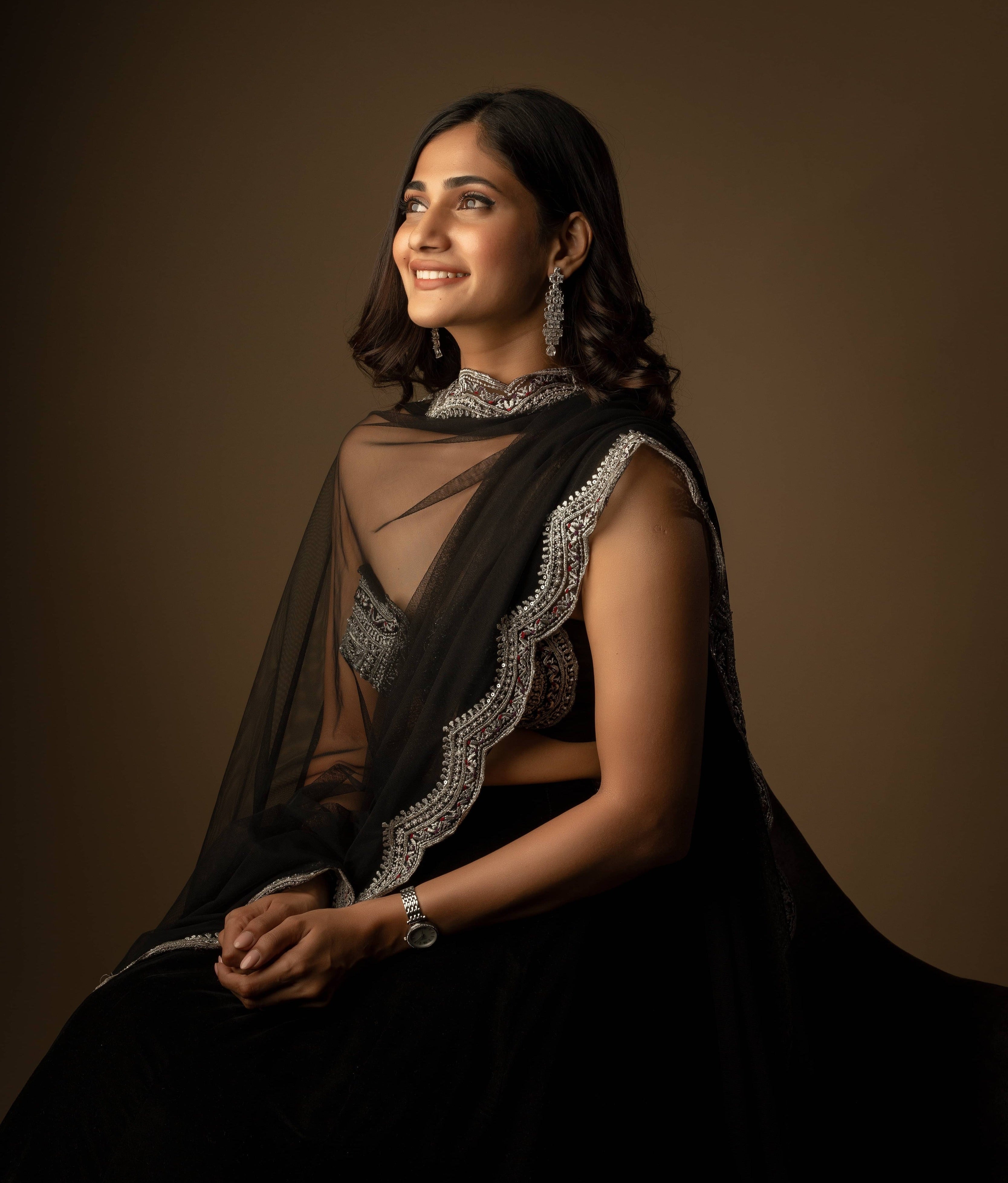 Bigg Boss fame and actress Losliya Mariyanesan in black velvet lehenga with silver embellishments on the borders and bralette. Seated pose looking away from camera