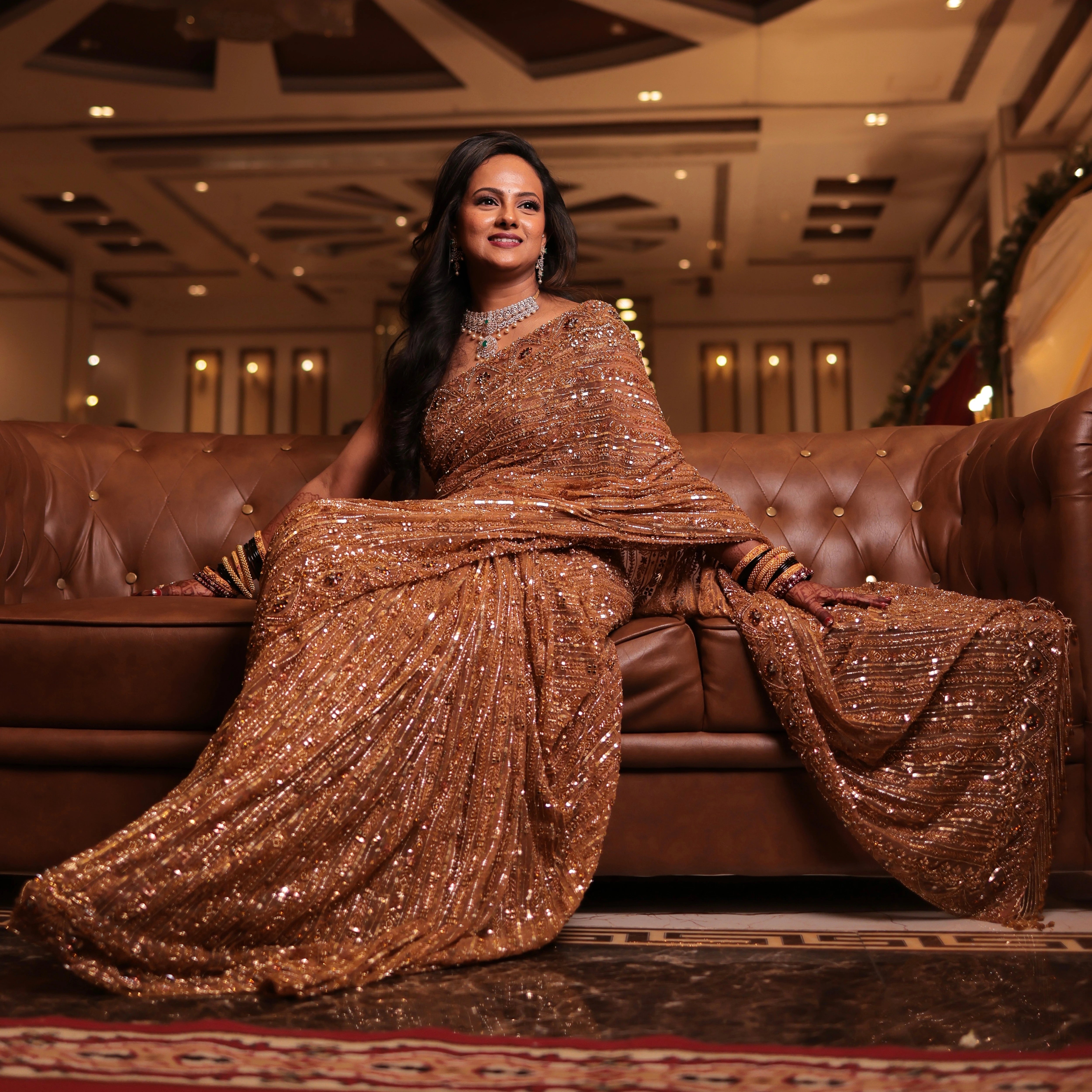 Bride Aishwarya from USA, posing on a sofa in a golden tulle saree handcrafted by team Archana Karthick.