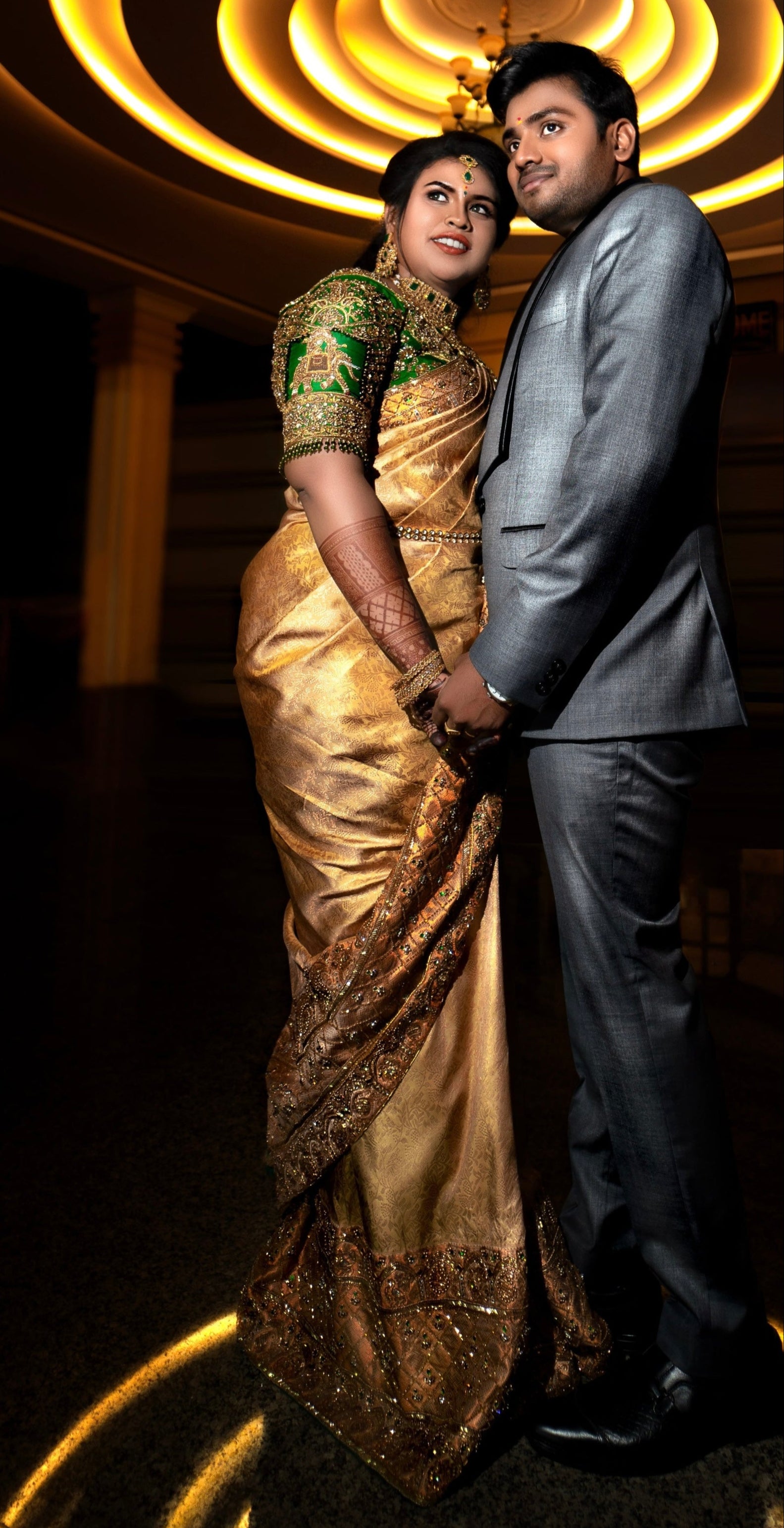 Bride Hemakiruthika from Chennai in a green raw silk blouse and gold silk saree with heavy border embellishments, designed by Archana Karthick. She is looking to her right and is posing with the groom.
