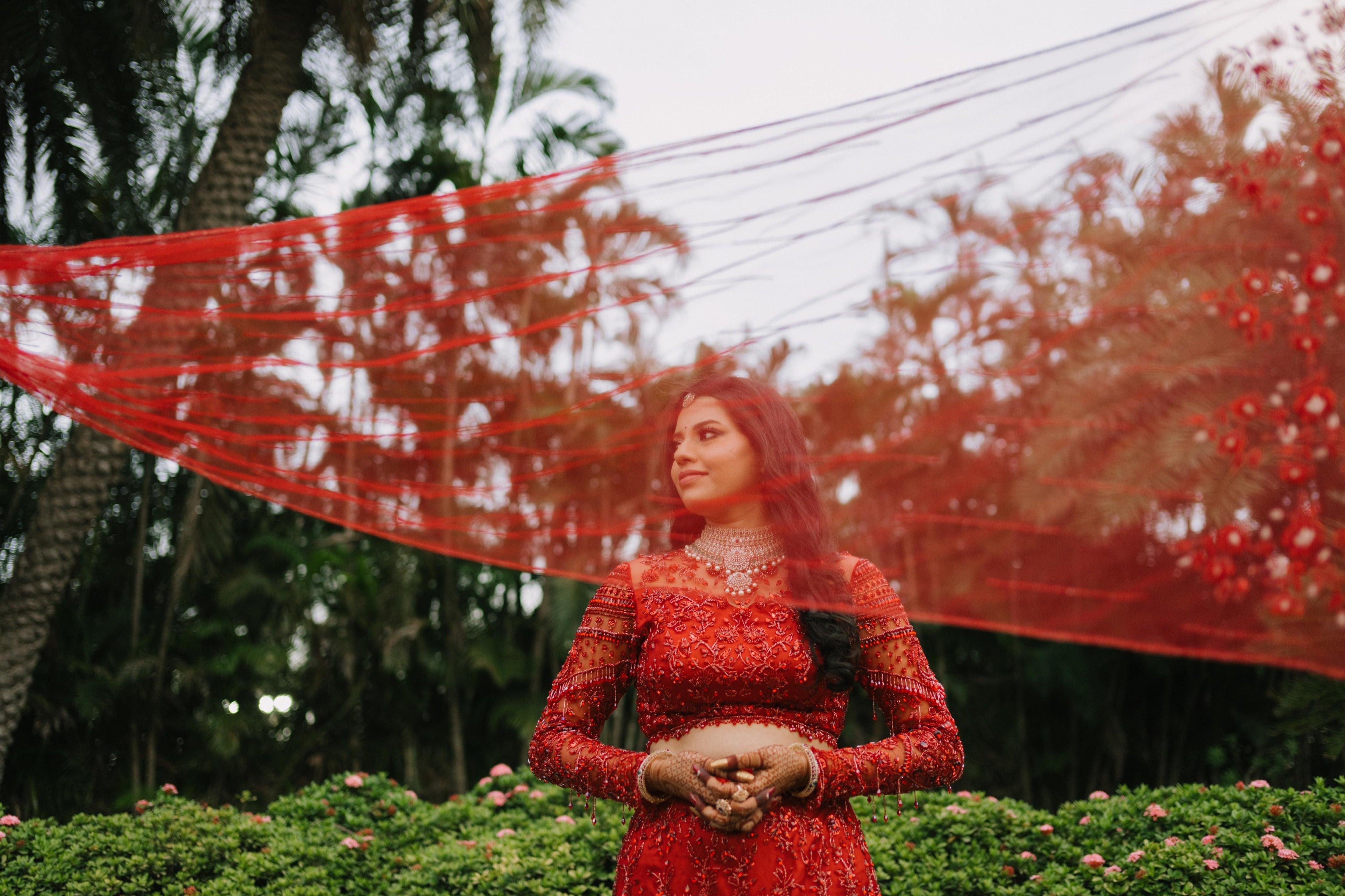 Bride Urmila posing elegantly with her detachable trail as a veil for her reception in a red handcrafted mermaid lehenga drape designed by Archana Karthick.