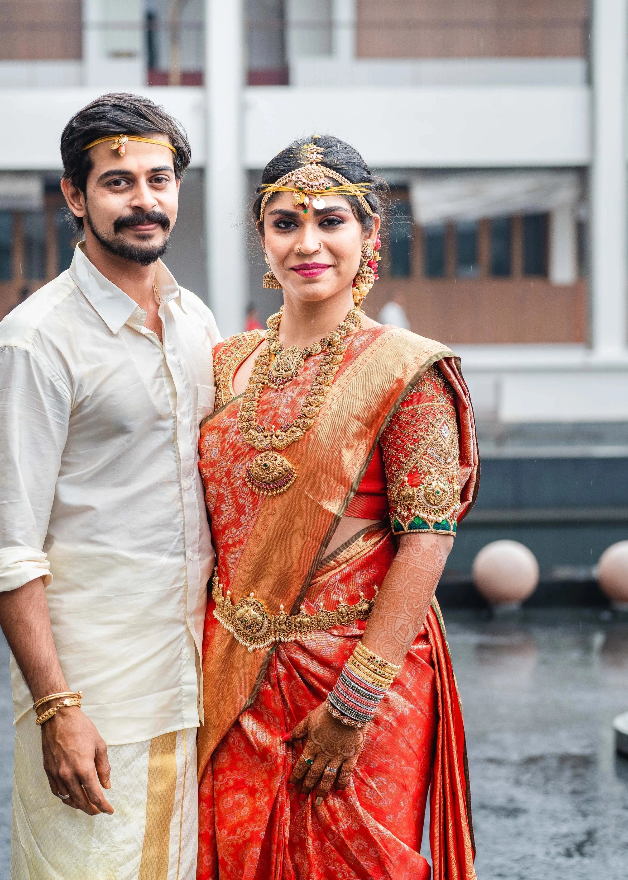 Bride Vidya D posing with groom in an orange silk blouse, handcrafted with beads, zardozi, kundan, resham, stones and zardozi by team Archana Karthick.