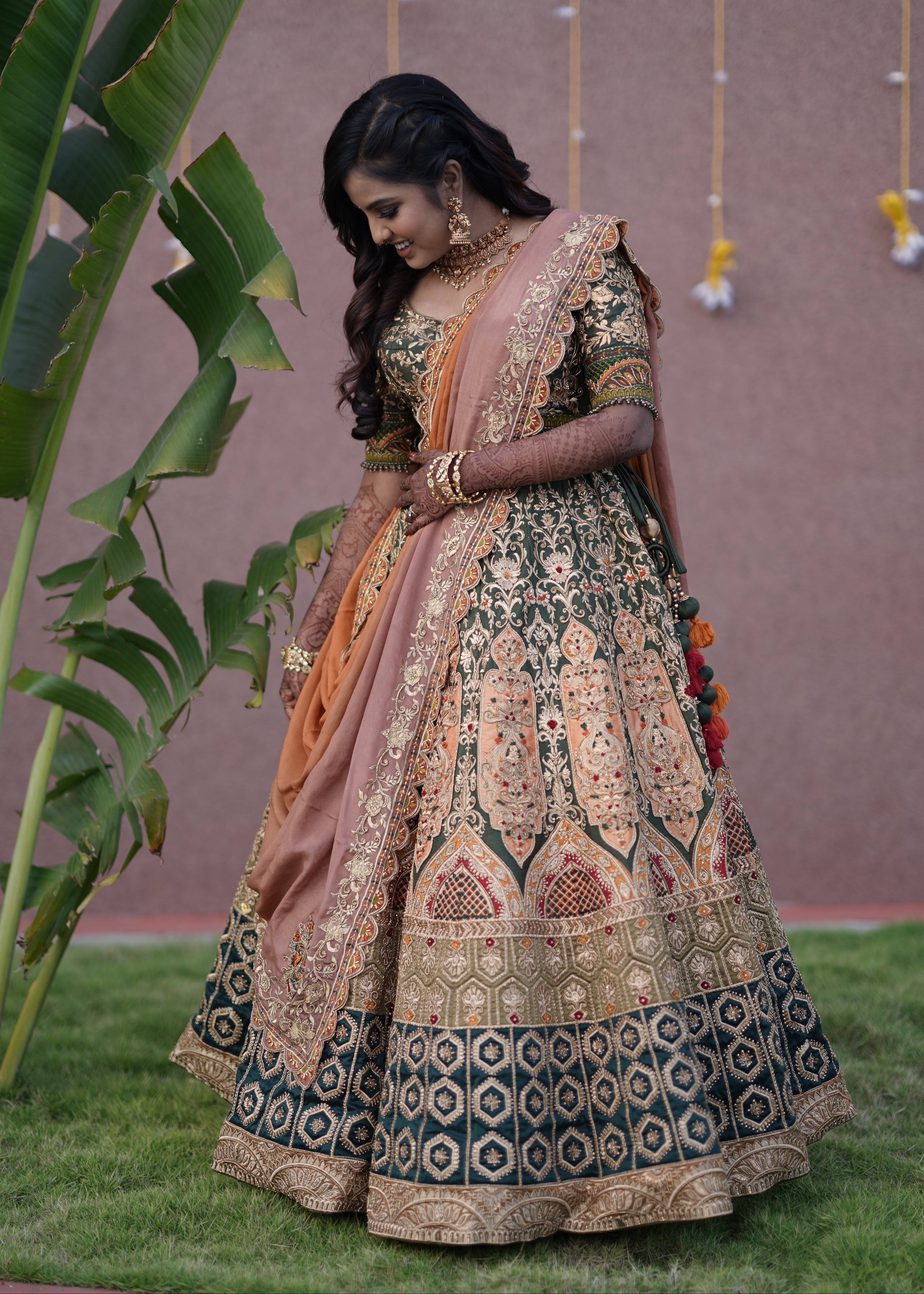 Bride from Chennai looking down in an olive green raw silk lehenga with hints of burnt orange handcrafted intricately with zardozi, crystal beads, resham knot work, cutdanas and sequins. Designed by Archana Karthick.