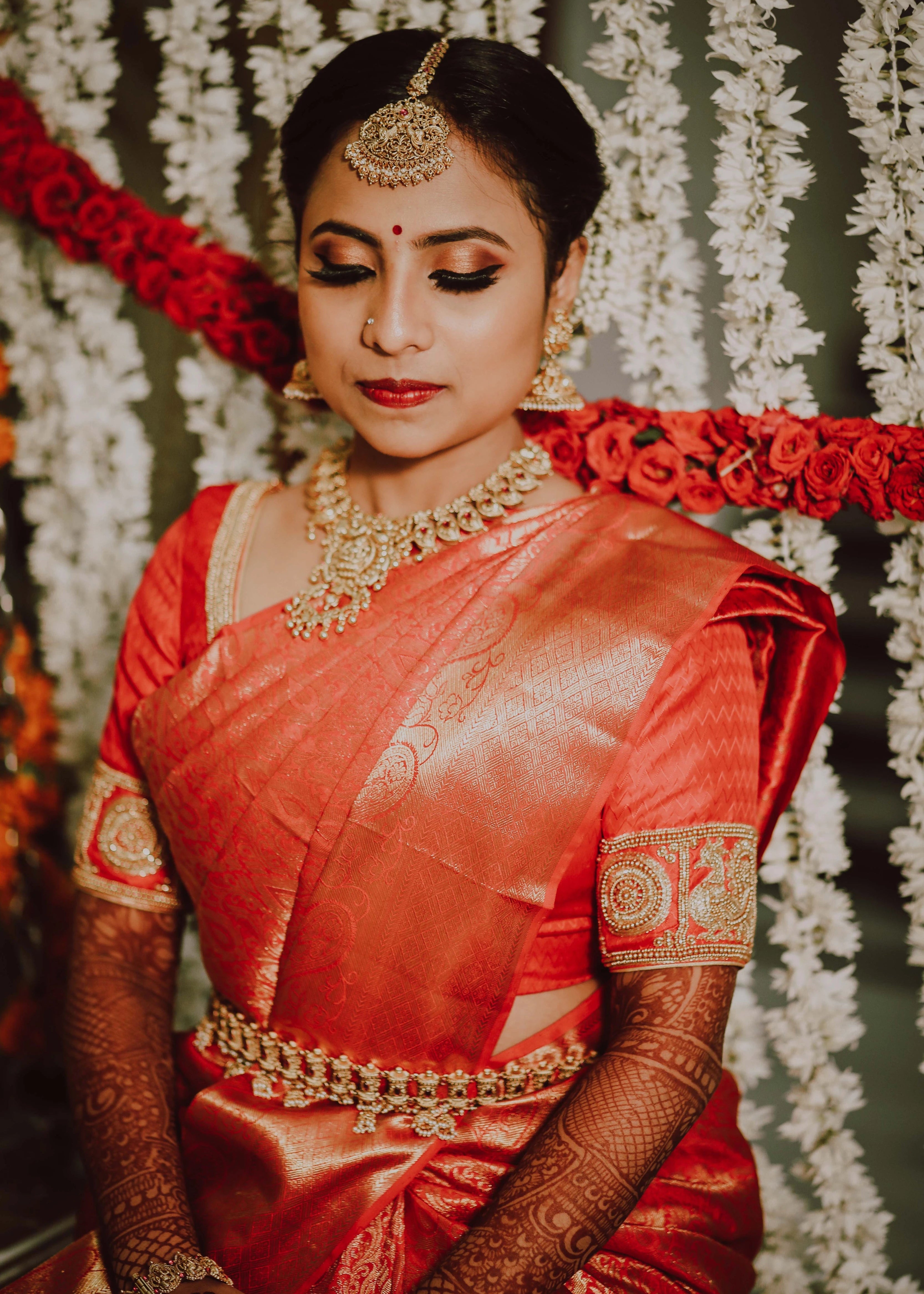 Bride from USA looking down in a red silk blouse for her engagement ceremony. Handcrafted intricately with beads, zardozi and sequins by team Archana Karthick.