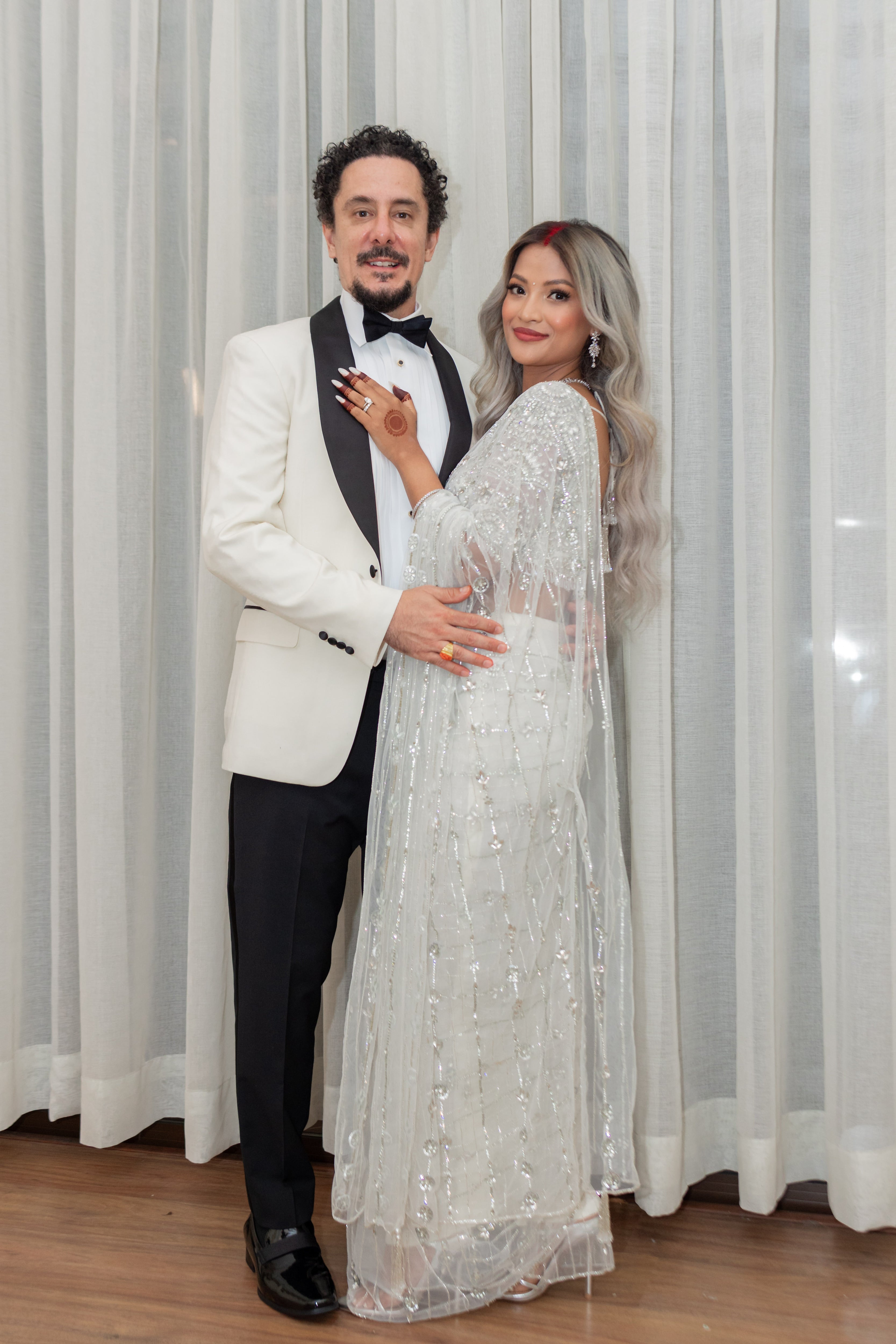 Bride from texas in white net saree for wedding reception in nepal. She is posing with the groom giving us a full side view of the lovely white saree. The saree shines with intricate stonework, sequins and cut dana details.