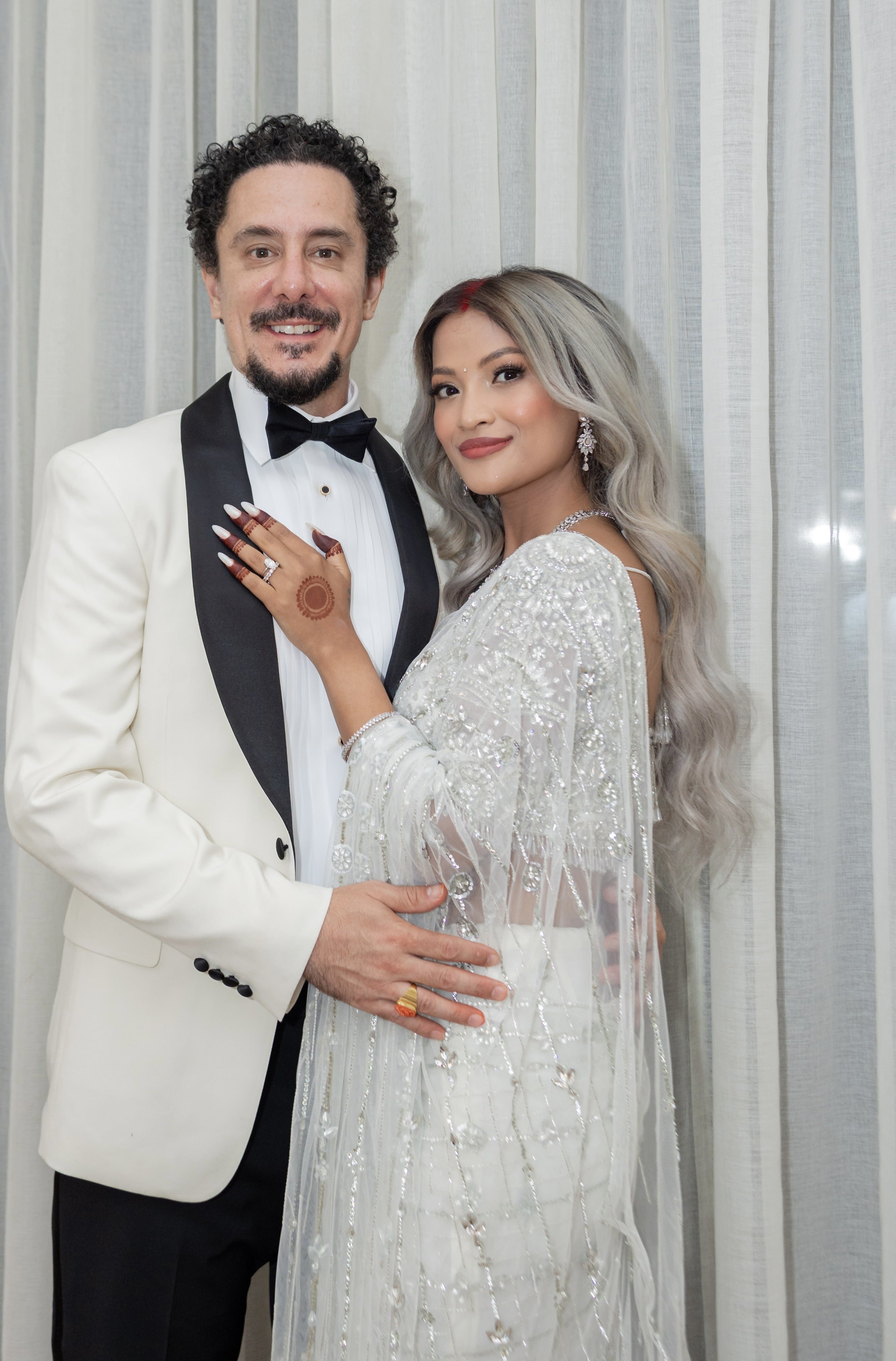 Bride from texas in white net saree for wedding reception in nepal. She is posing with the groom giving us a half side view of the lovely white saree. The saree shines with intricate stonework, sequins and cut dana details.