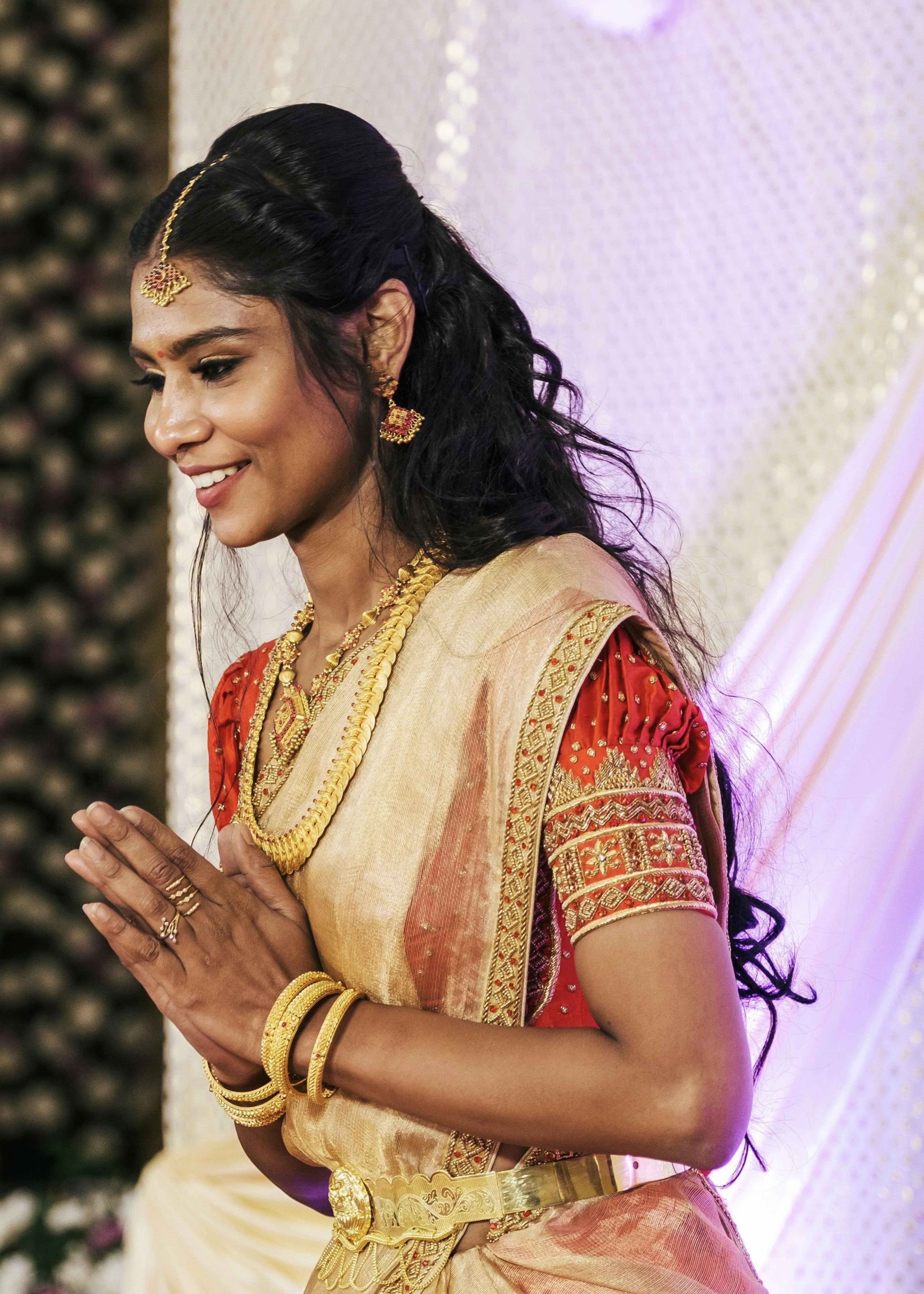 Bride greeting guests in a handcrafted red silk half saree with katori cut blouse has intricate zardozi and stone work.