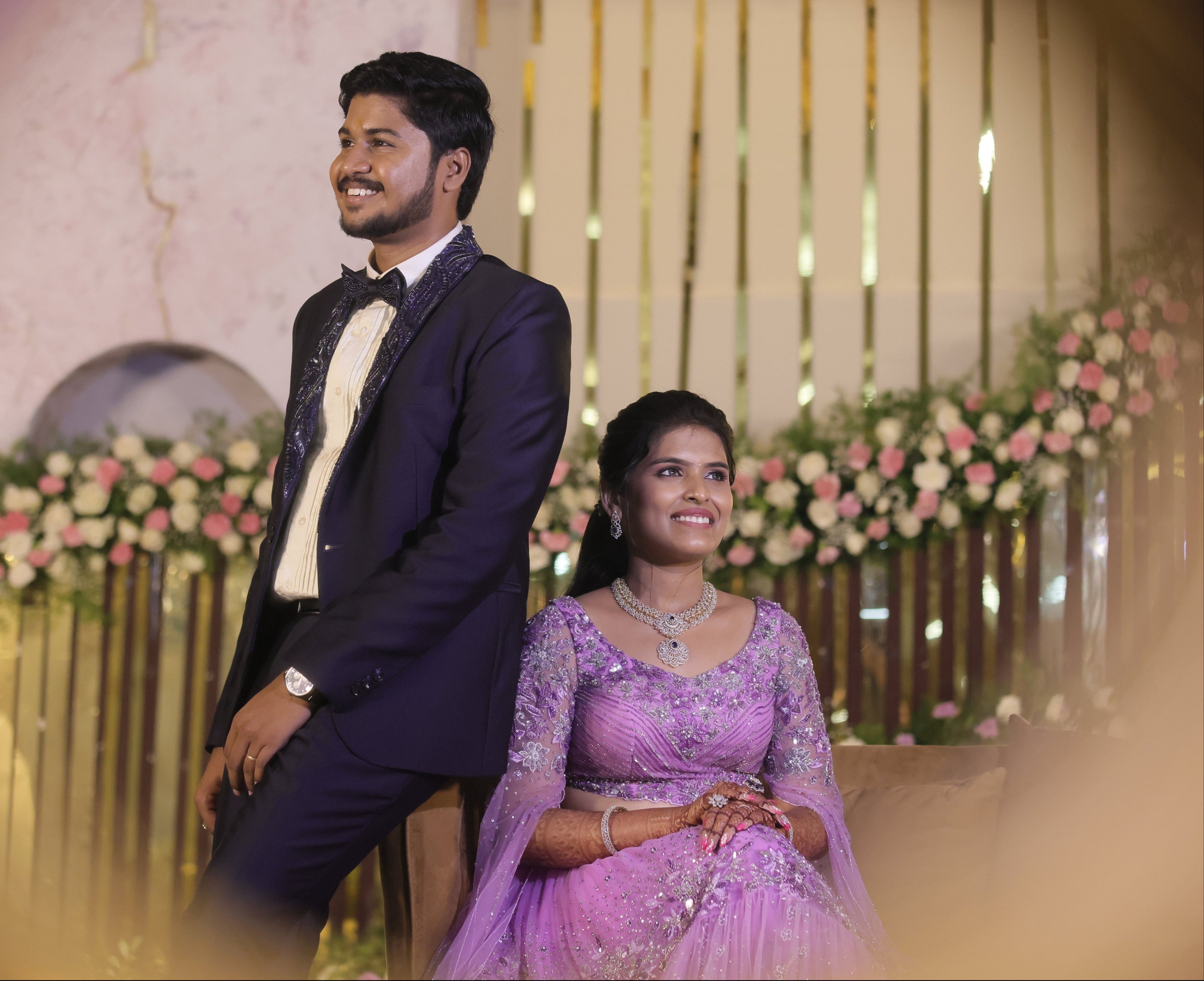 Bride in a lavender handcrafted lehenga with trail sleeves and groom in a bespoke navy two piece suit with handcrafted lapels and bow tie for their reception. The outfits for both the bride and groom is designed by Archana Karthick. They are posing on the reception stage.