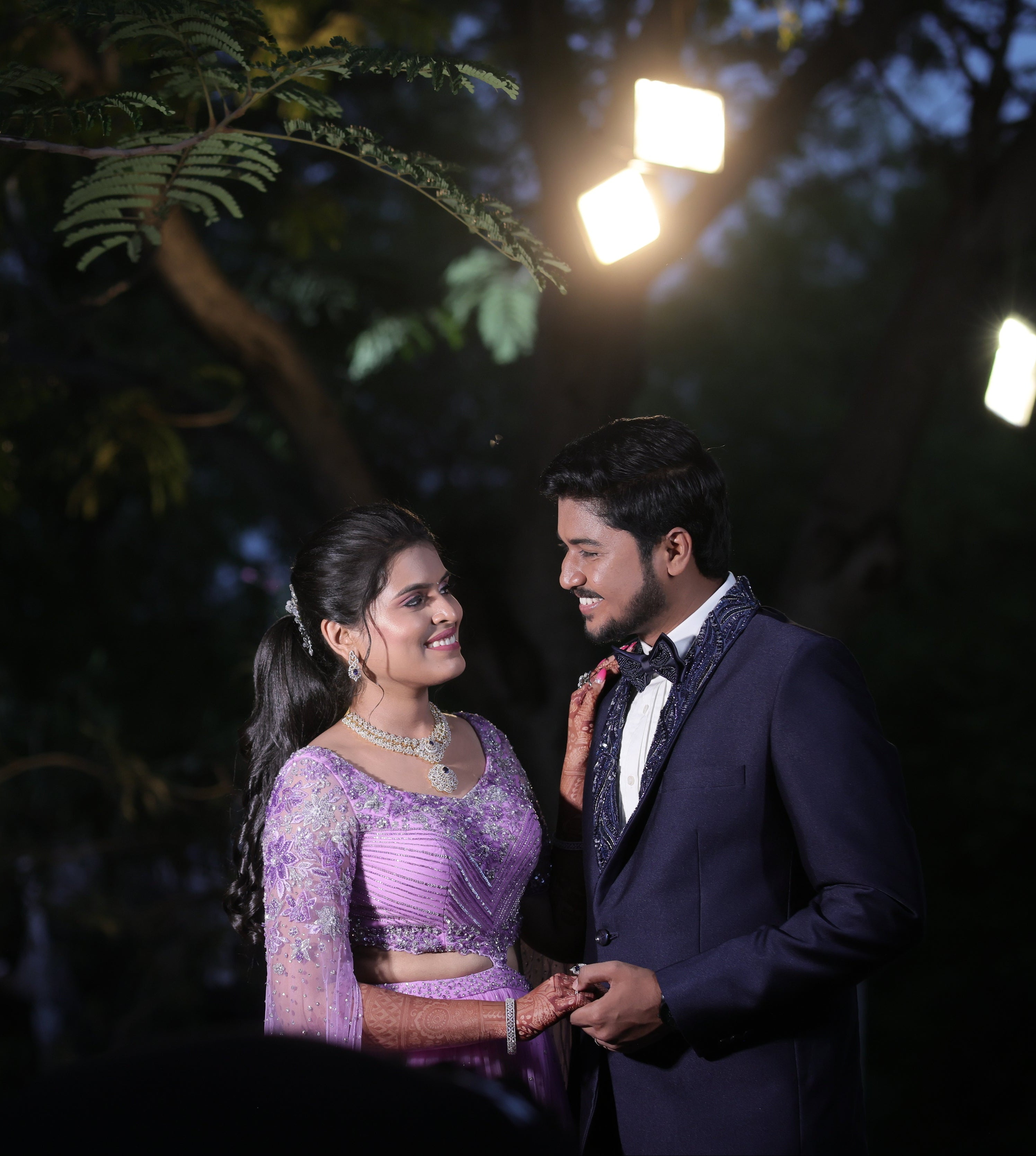 Bride in a lavender handcrafted lehenga with trail sleeves and groom in a bespoke navy two piece suit with handcrafted lapels and bow tie for their reception. The outfits for both the bride and groom is designed by Archana Karthick. They are smiling at each other.