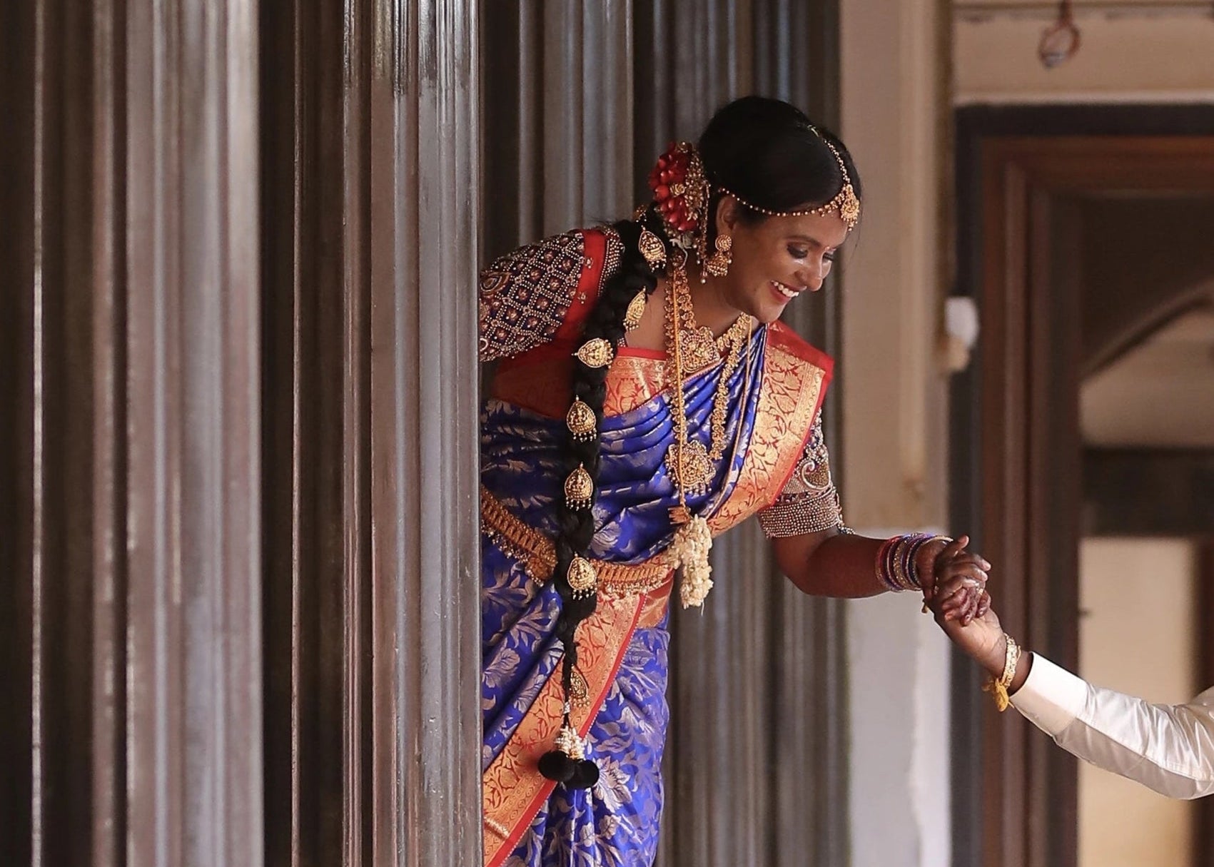 Bride looking down for photoshoot in a concept peacock blouse in blue and red silk, handcrafted with zardozi, beads, resham knot work and goddess pendant on sleeves.