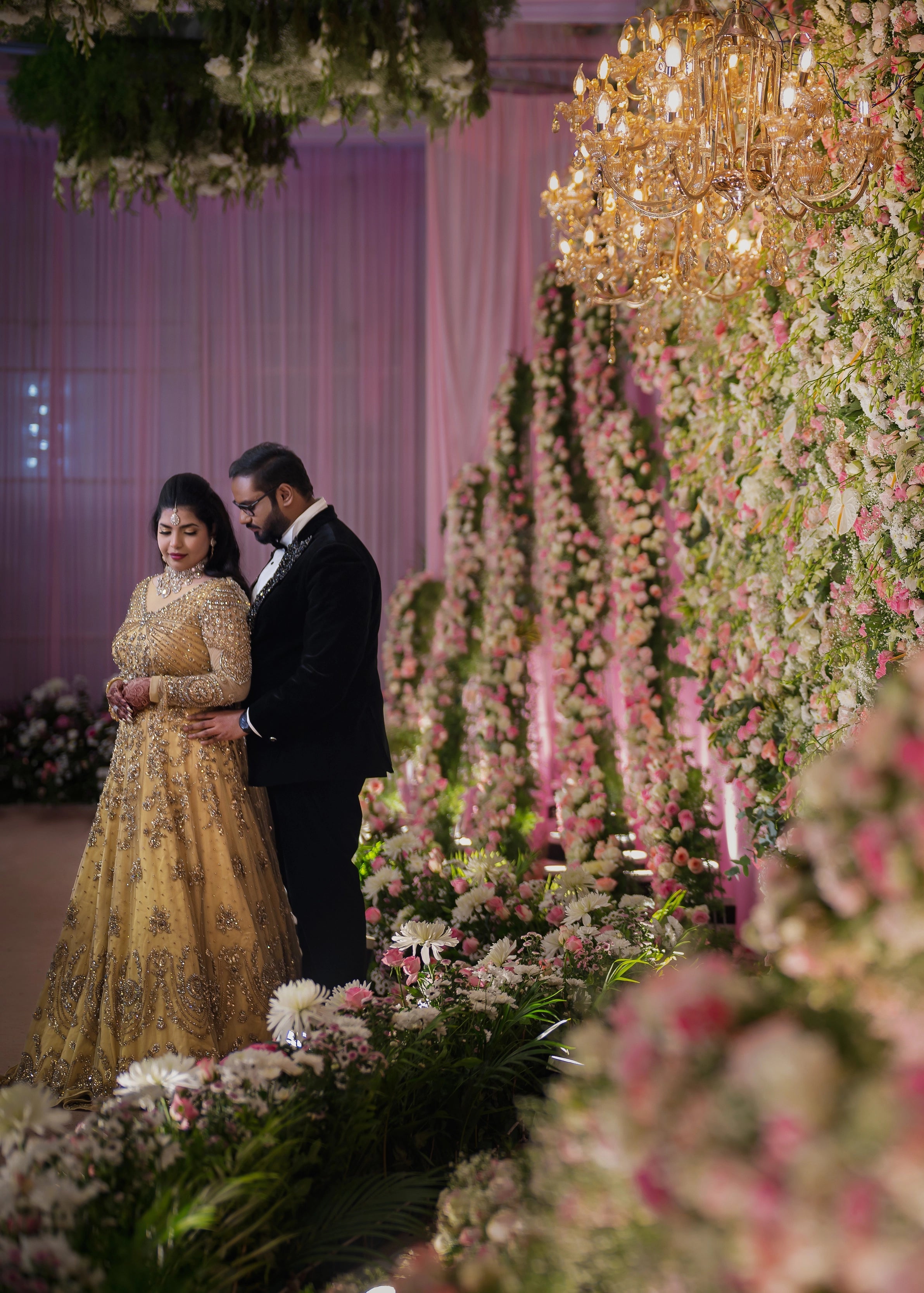 Bride posing with groom on stage in a gold gown in tulle designed by Archana Karthick and handcrafted with rhinestones and cut dana with floral and geometric motifs.