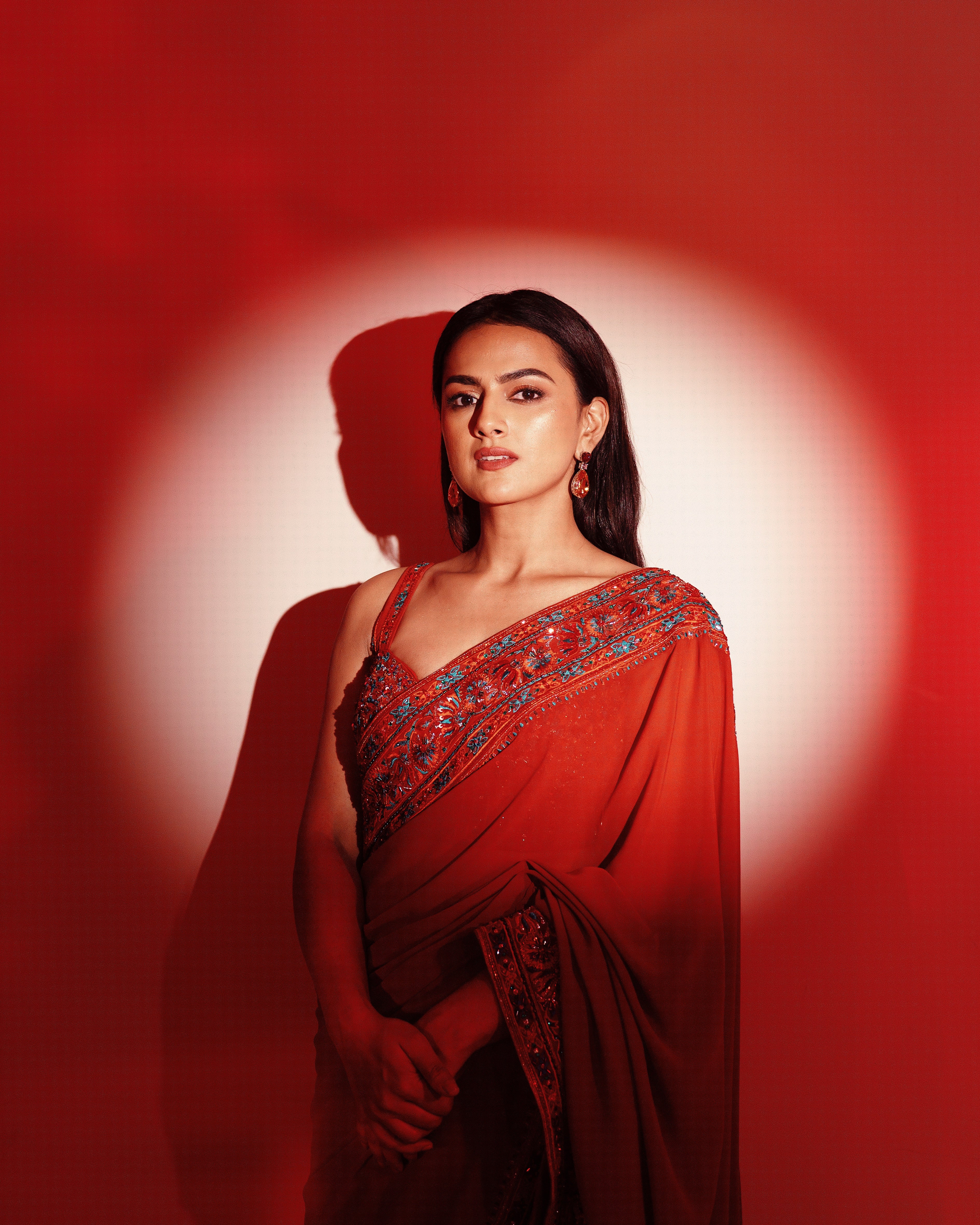 Indian film actress Shraddha Srinath poses in a burnt orange saree by Archana Karthick, featuring intricate embroidery on the blouse and borders, against a bright orange backdrop.