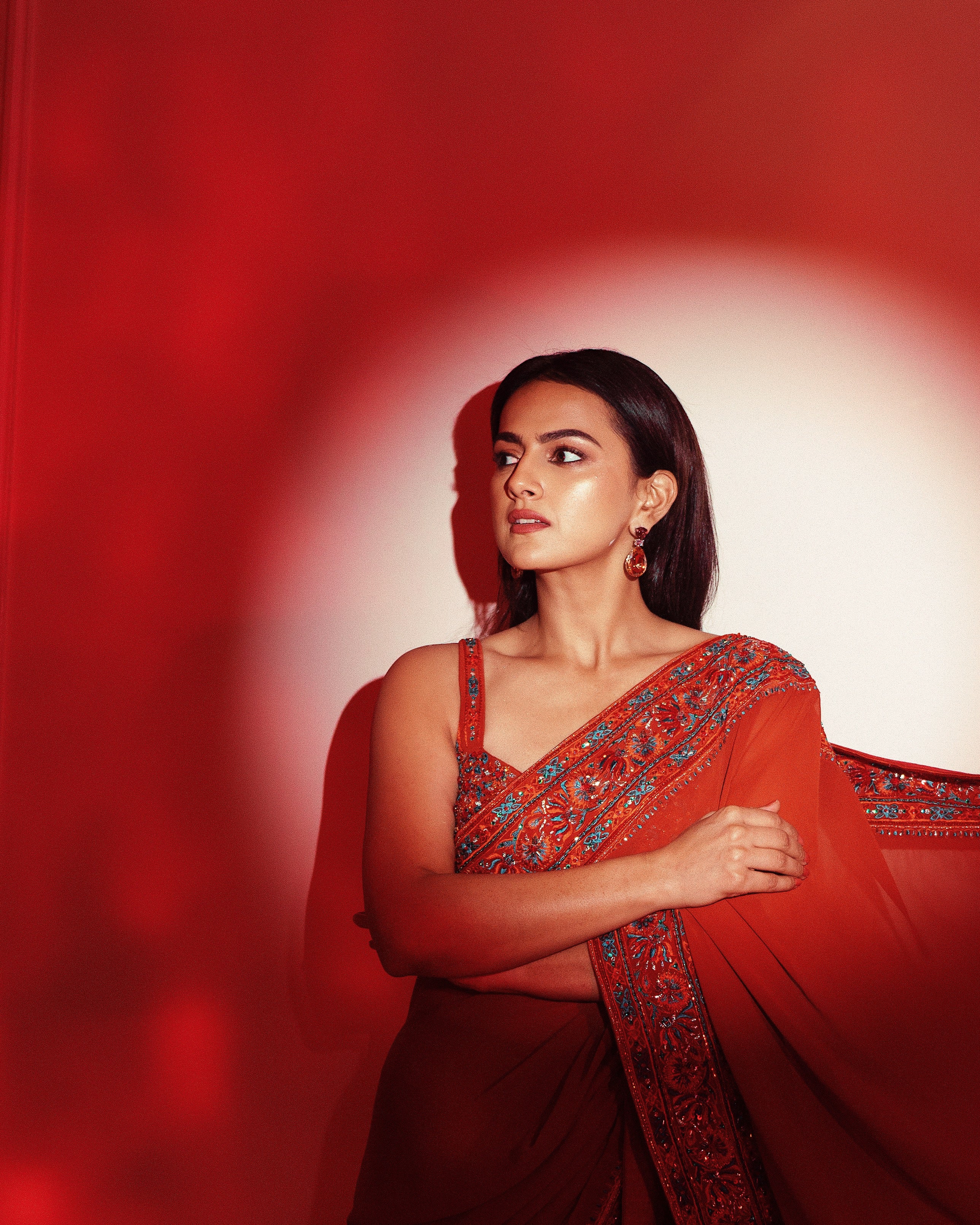 Indian film actress Shraddha Srinath poses in a burnt orange saree by Archana Karthick, featuring intricate embroidery on the blouse and borders, against the backdrop of a an orange glow.