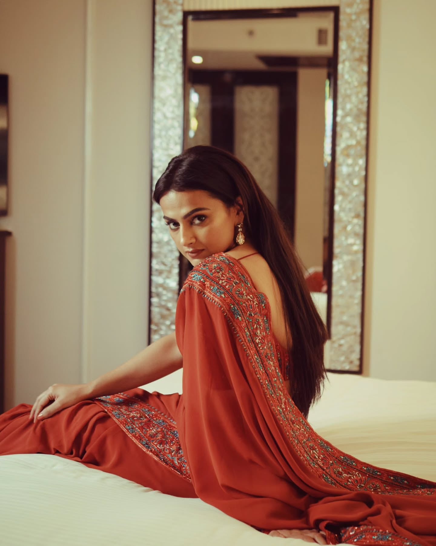 Indian film actress Shraddha Srinath poses in a burnt orange saree by Archana Karthick, featuring intricate embroidery on the blouse and borders, against the backdrop of a mirror.