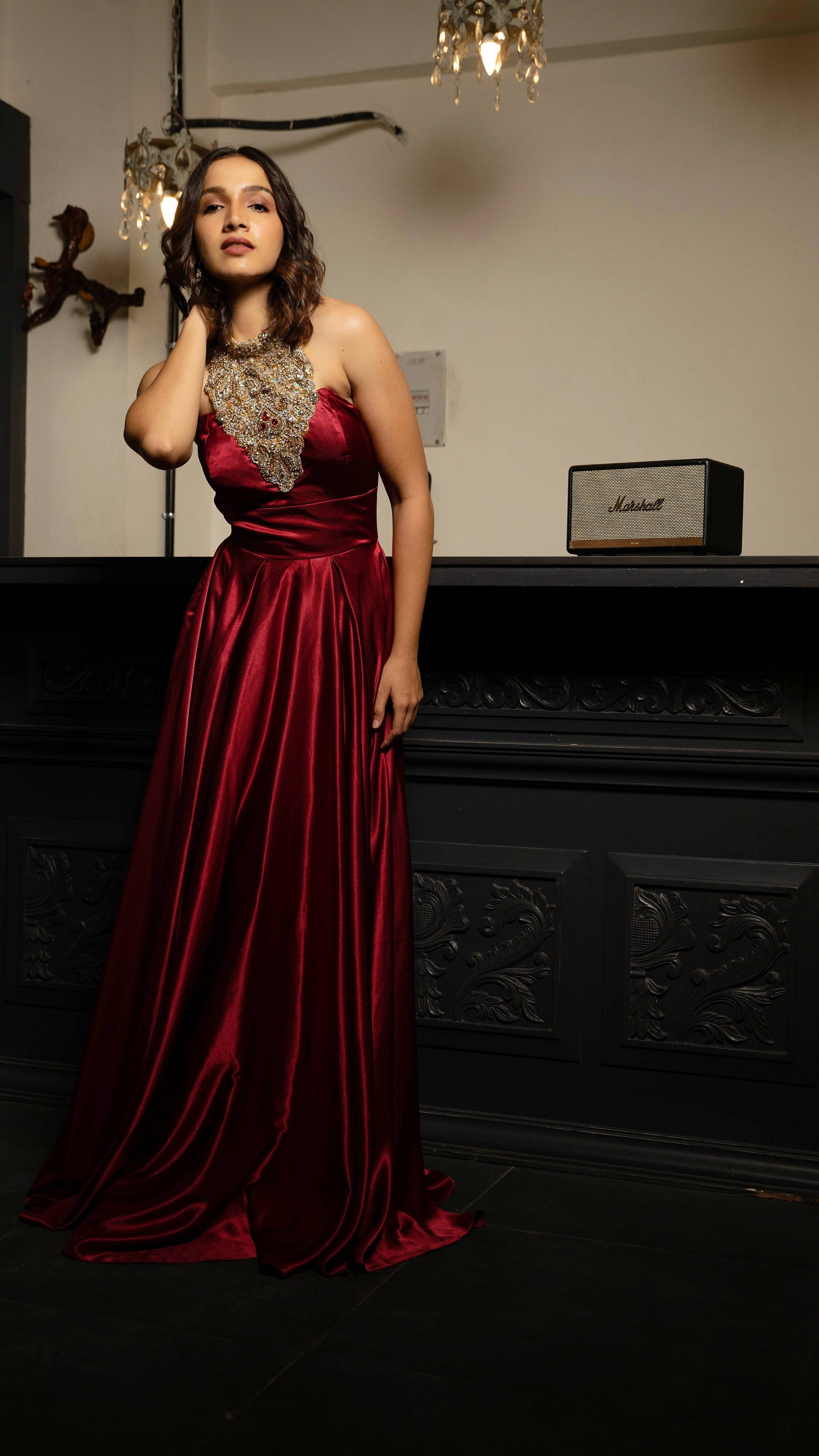 Influencer and actress Shweta Gai in a red satin silk halterneck gown with a heavily embellished neckline. Bent forward and looking at the camera.