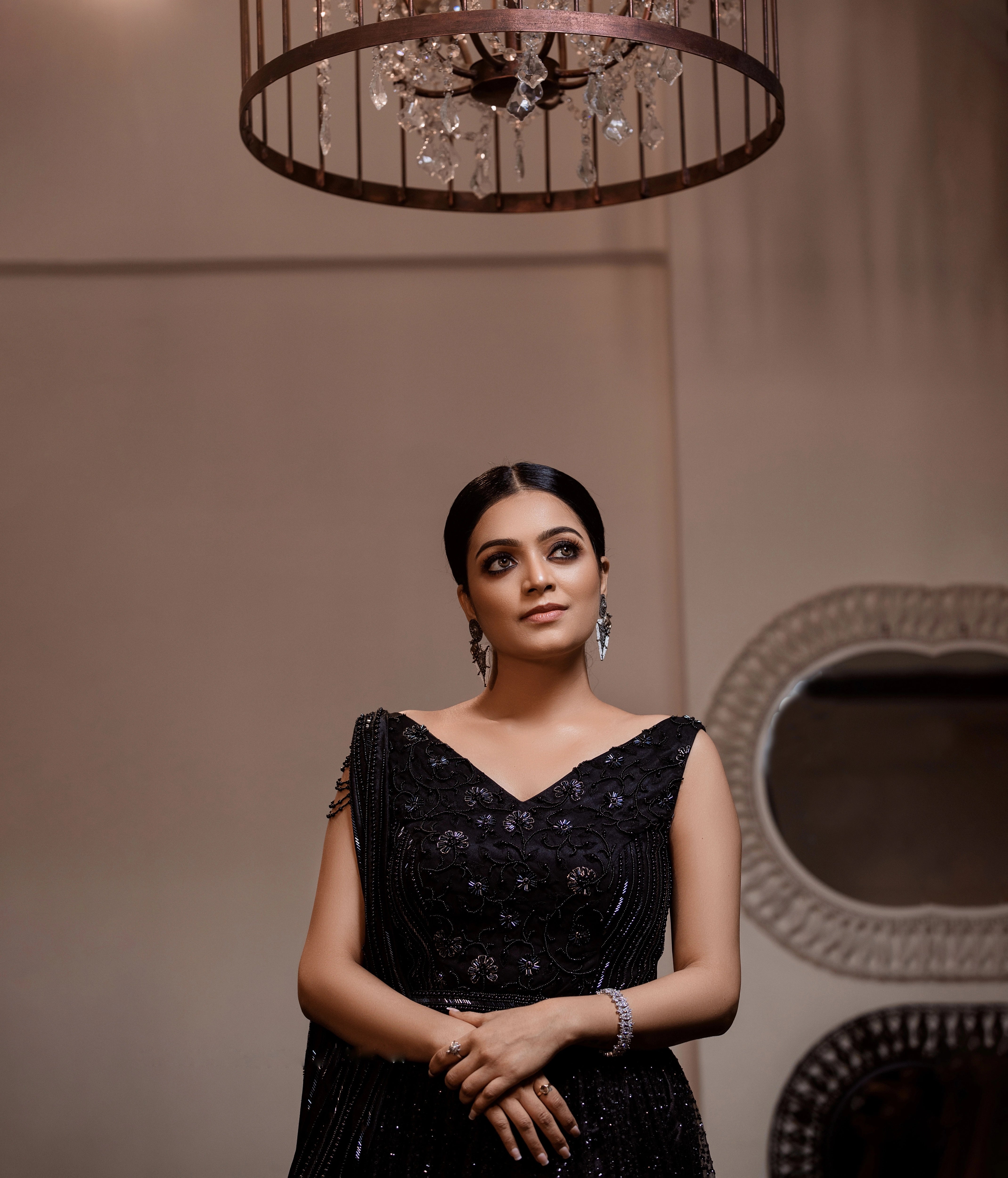 Janani Iyer posing under chandelier in a black net and satin gown with handcrafted drape, intricately designed with cutdana, sequins and beads by team Archana Karthick.