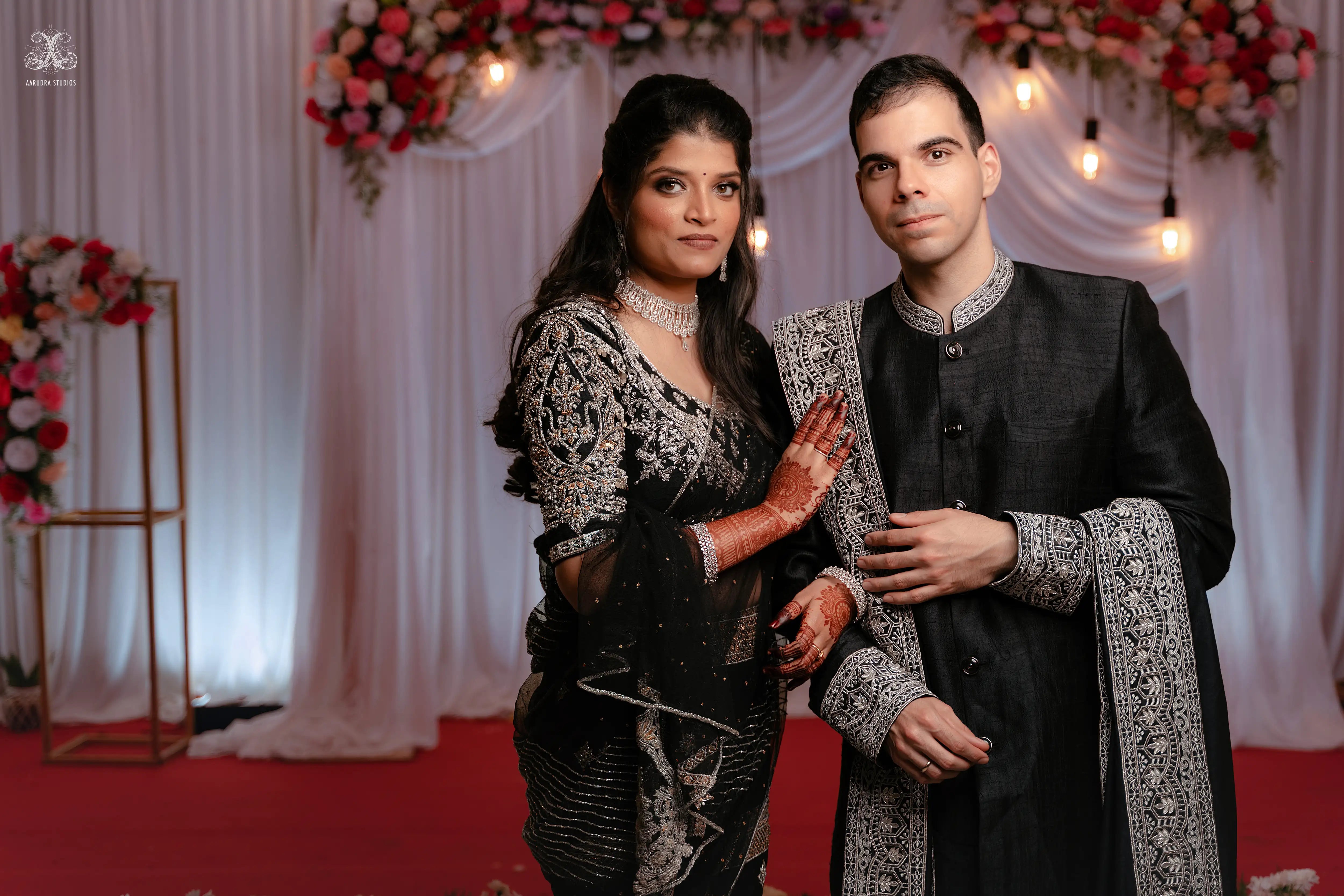 Kamakshi & Alex from Yokosuka, Japan, posing on stage in a black raw silk lehenga and sherwani handcrafted by team Archana Karthick.
