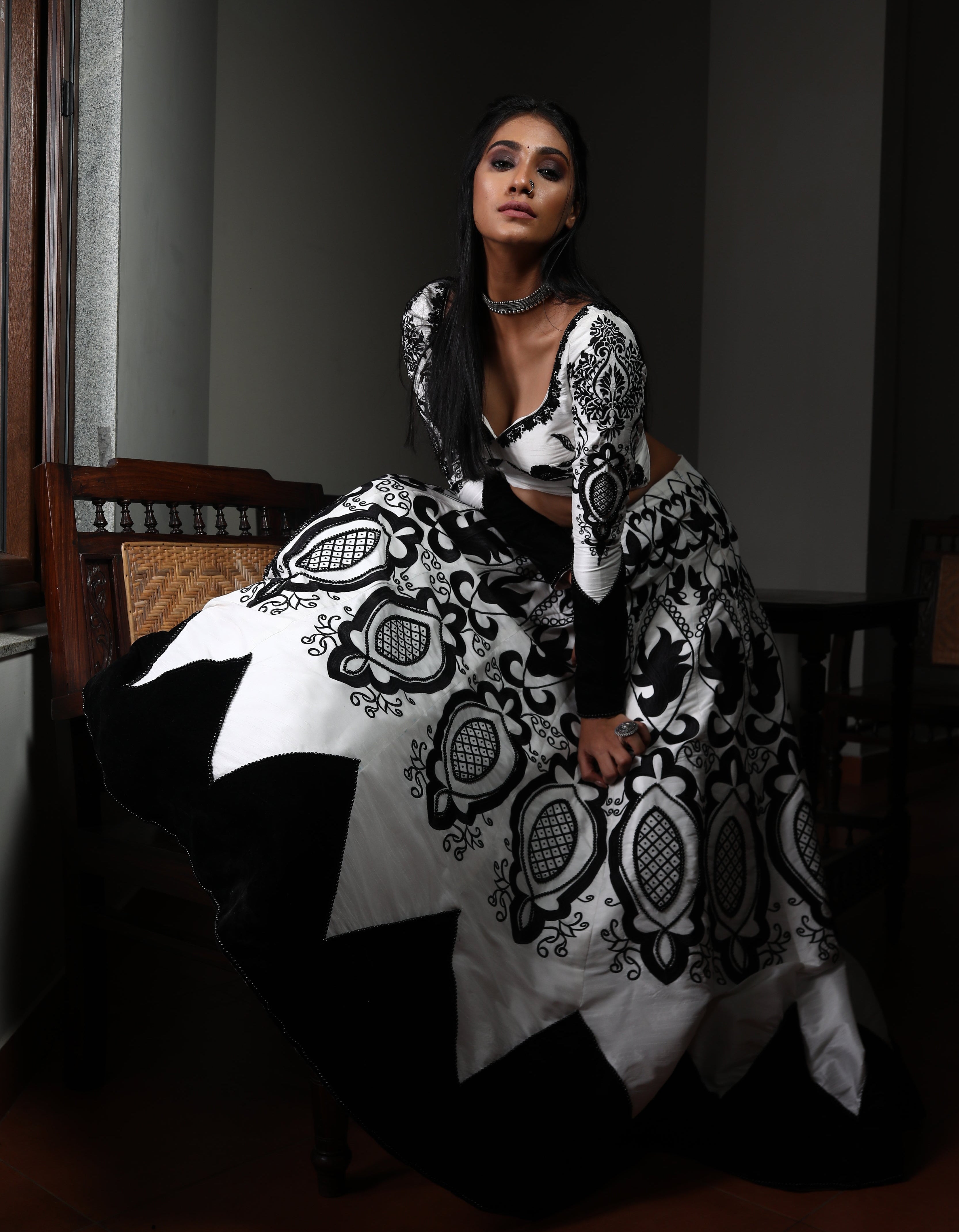 Miss India 2018 Anukreethy Vas in white raw sIlk lehenga with motifs in black velvet posing bent forward and looking straight at the lens with one leg on the antique sofa. This lehenga is designed by Archana Karthick