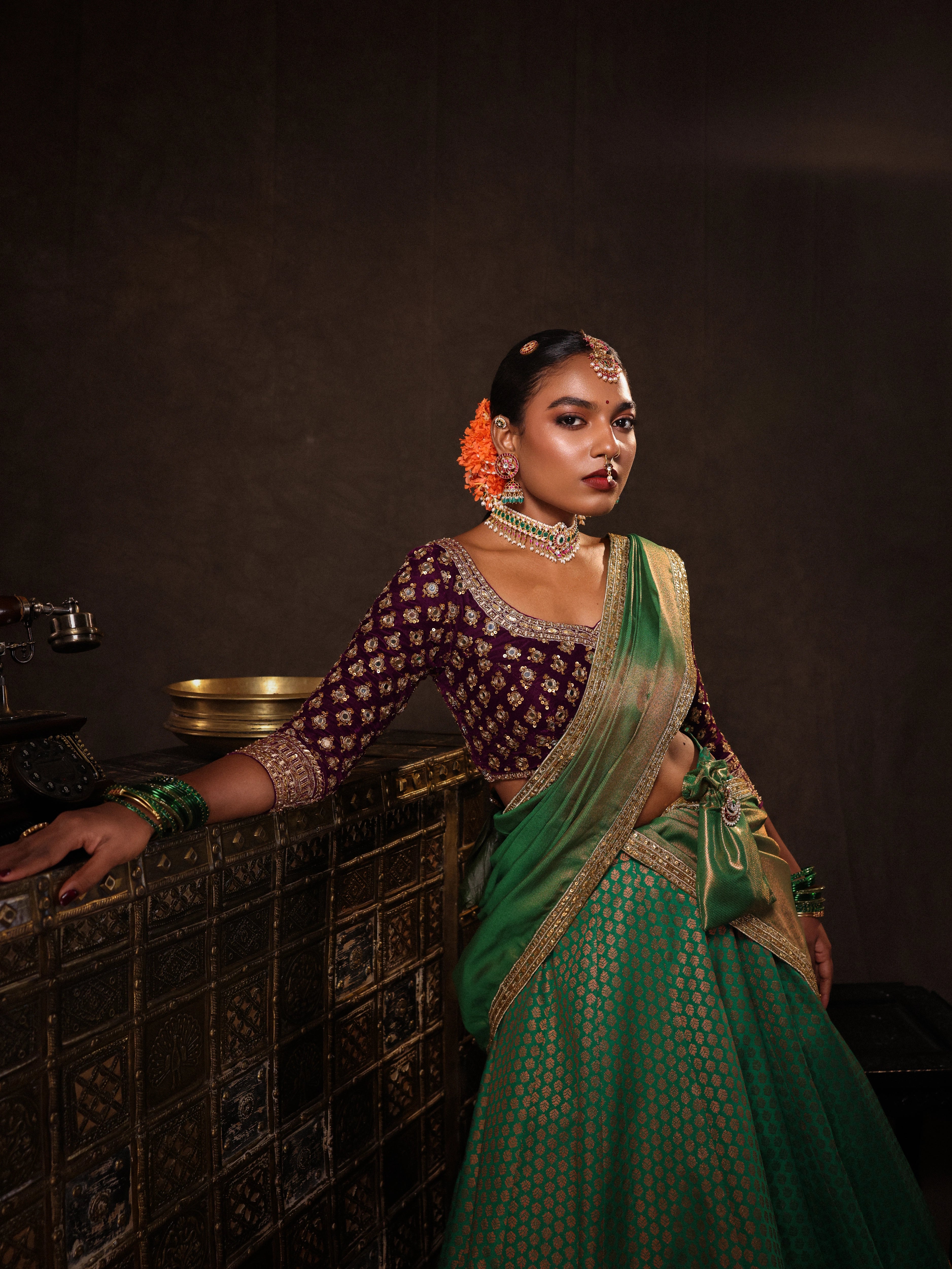 Model Ashika Crystal in a leaning pose in a green and purple half saree set with a velvet blouse, tissue silk dupatta and brocade skirt handcrafted with zardozi, mirror, zari, cutdana and sequins. Team Archana Karthick completed the style with green glass bangles, a ring, a choker, jhumkas, nose ring and a maang tikka.