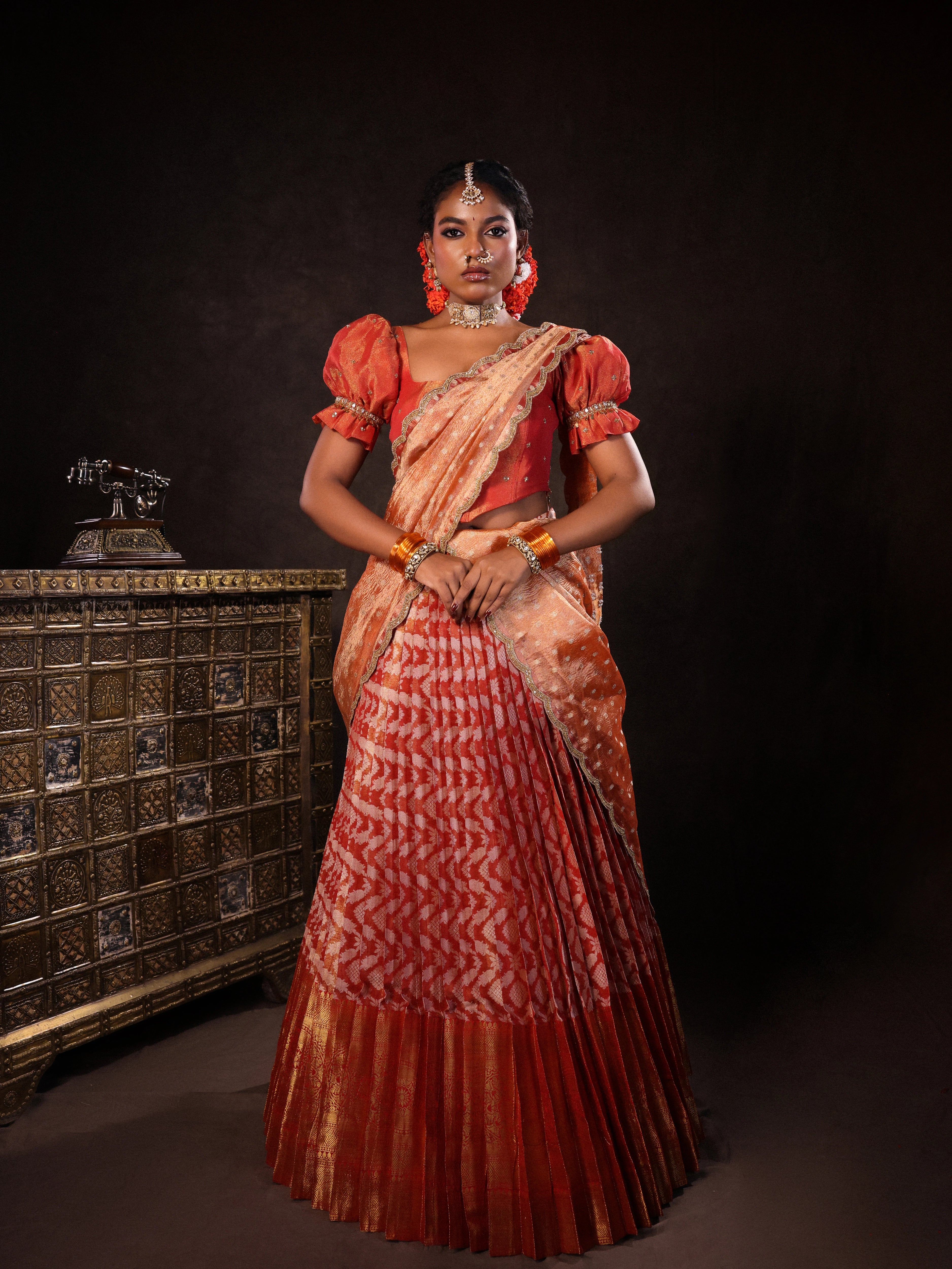 Model Ashika Crystal in a puff sleeved orange half saree set, handcrafted with zardozi, sequins, metal coin hangings and square cut stones. Team Archana Karthick completed the style with orange glass bangles, a ring, jhumkas, nose ring and a maang tikka.