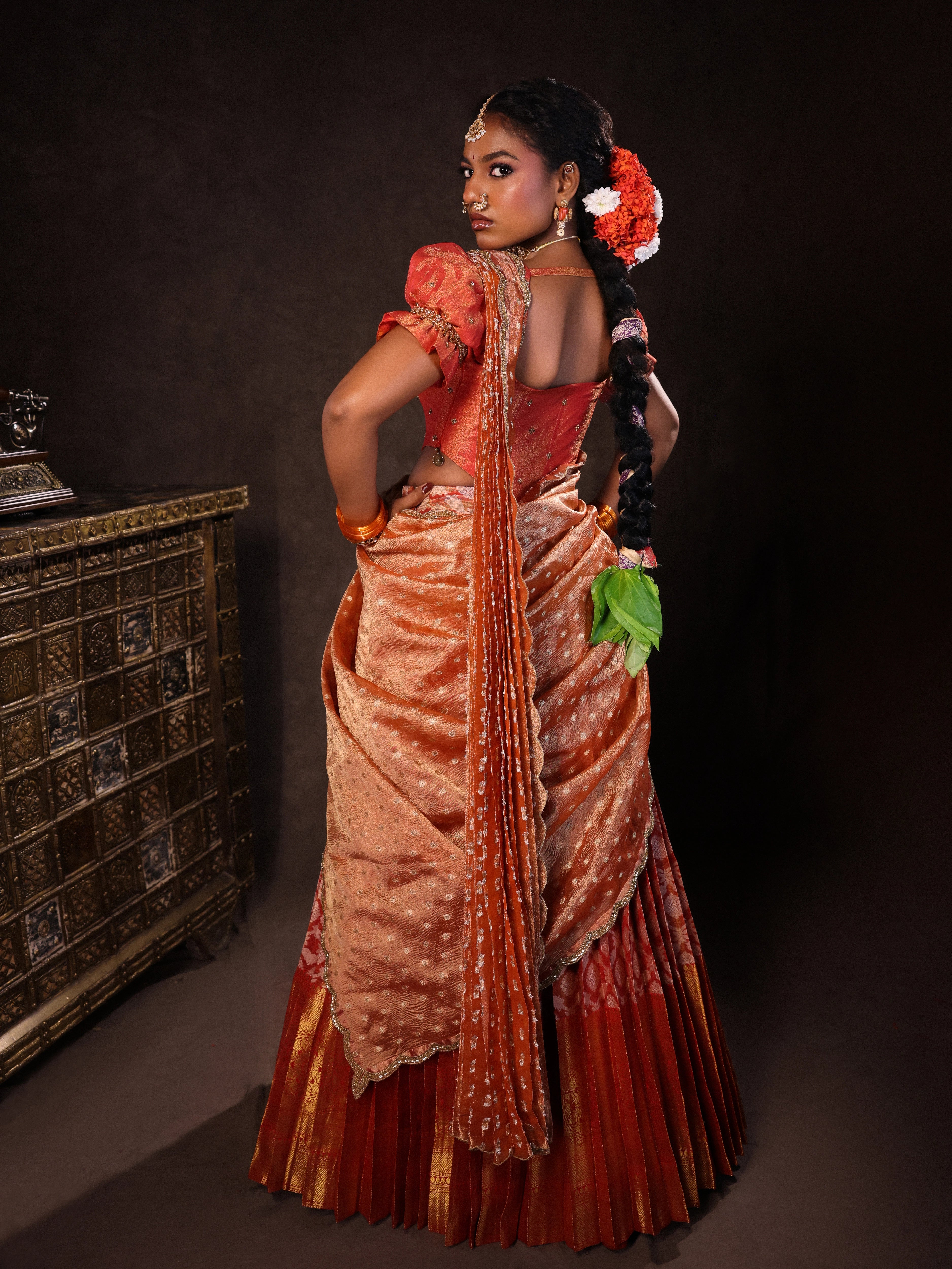 Model Ashika Crystal posing for back profile in a puff sleeved orange half saree set, handcrafted with zardozi, sequins, metal coin hangings and square cut stones. Team Archana Karthick completed the style with orange glass bangles, a ring, jhumkas, nose ring and a maang tikka.