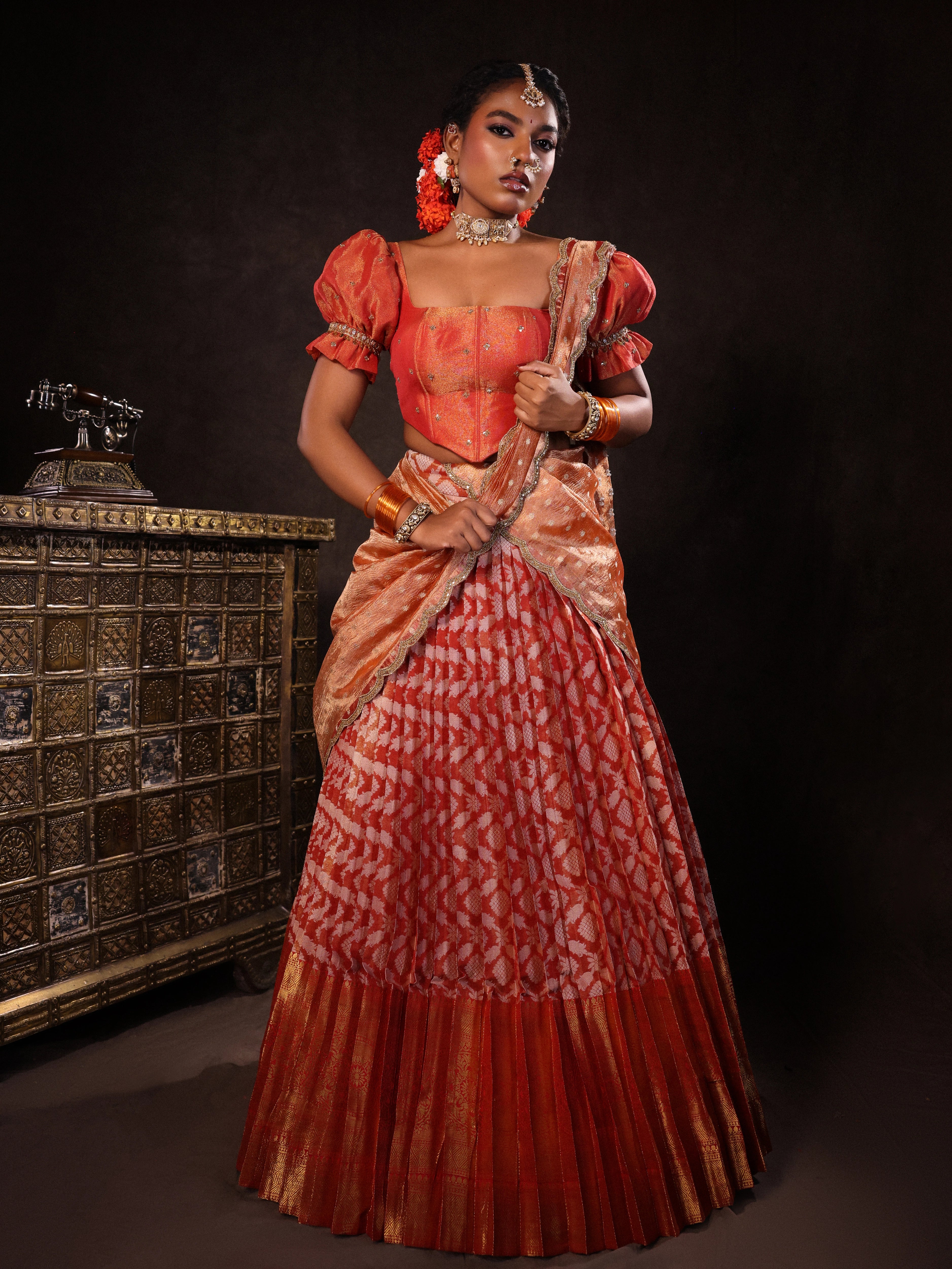 Model Ashika Crystal posing for front design in a puff sleeved orange half saree set, handcrafted with zardozi, sequins, metal coin hangings and square cut stones. Team Archana Karthick completed the style with orange glass bangles, a ring, jhumkas, nose ring and a maang tikka.