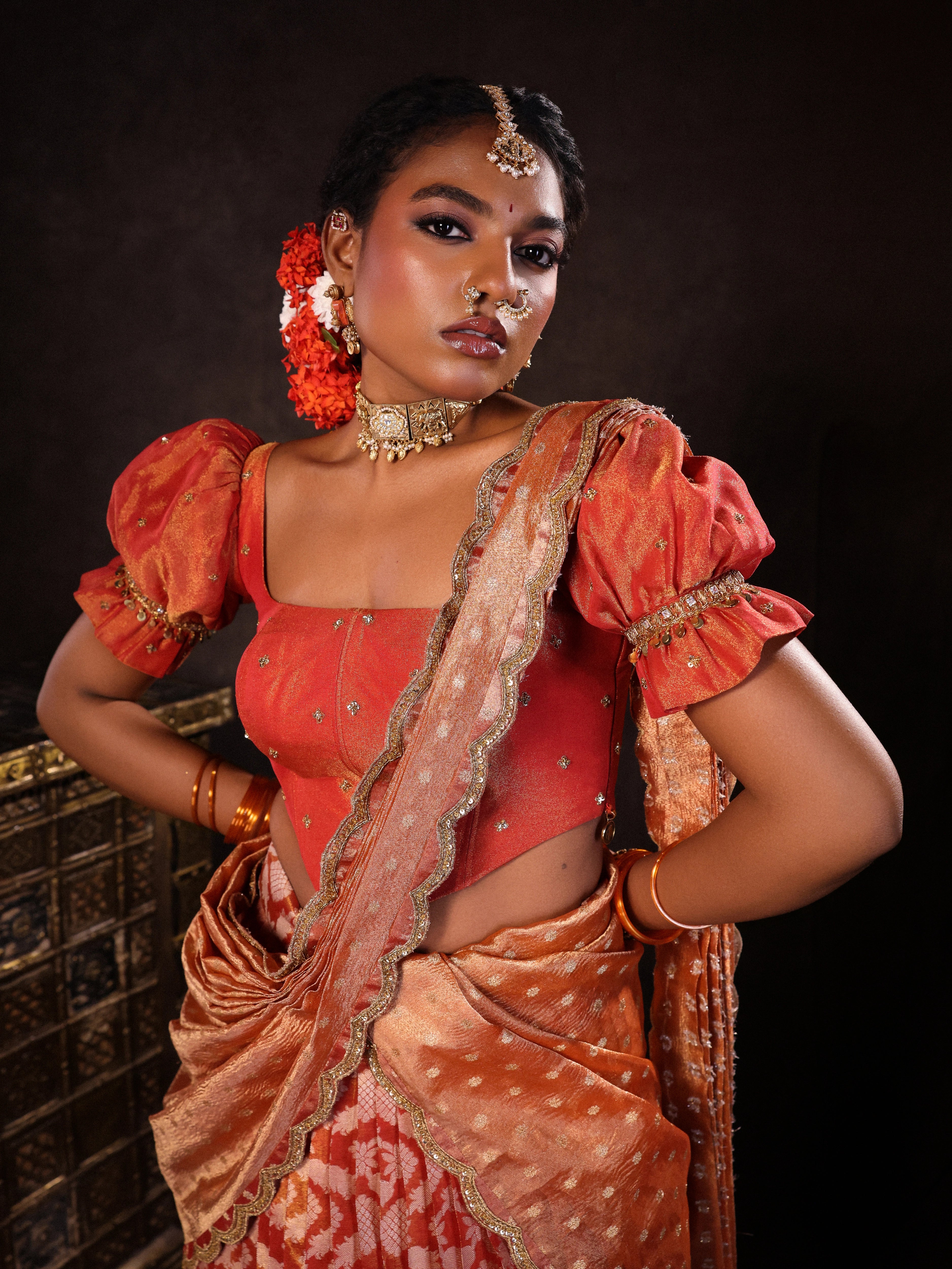 Model Ashika Crystal posing in a puff sleeved orange half saree set, handcrafted with zardozi, sequins, metal coin hangings and square cut stones. Team Archana Karthick completed the style with orange glass bangles, a ring, jhumkas, nose ring and a maang tikka.
