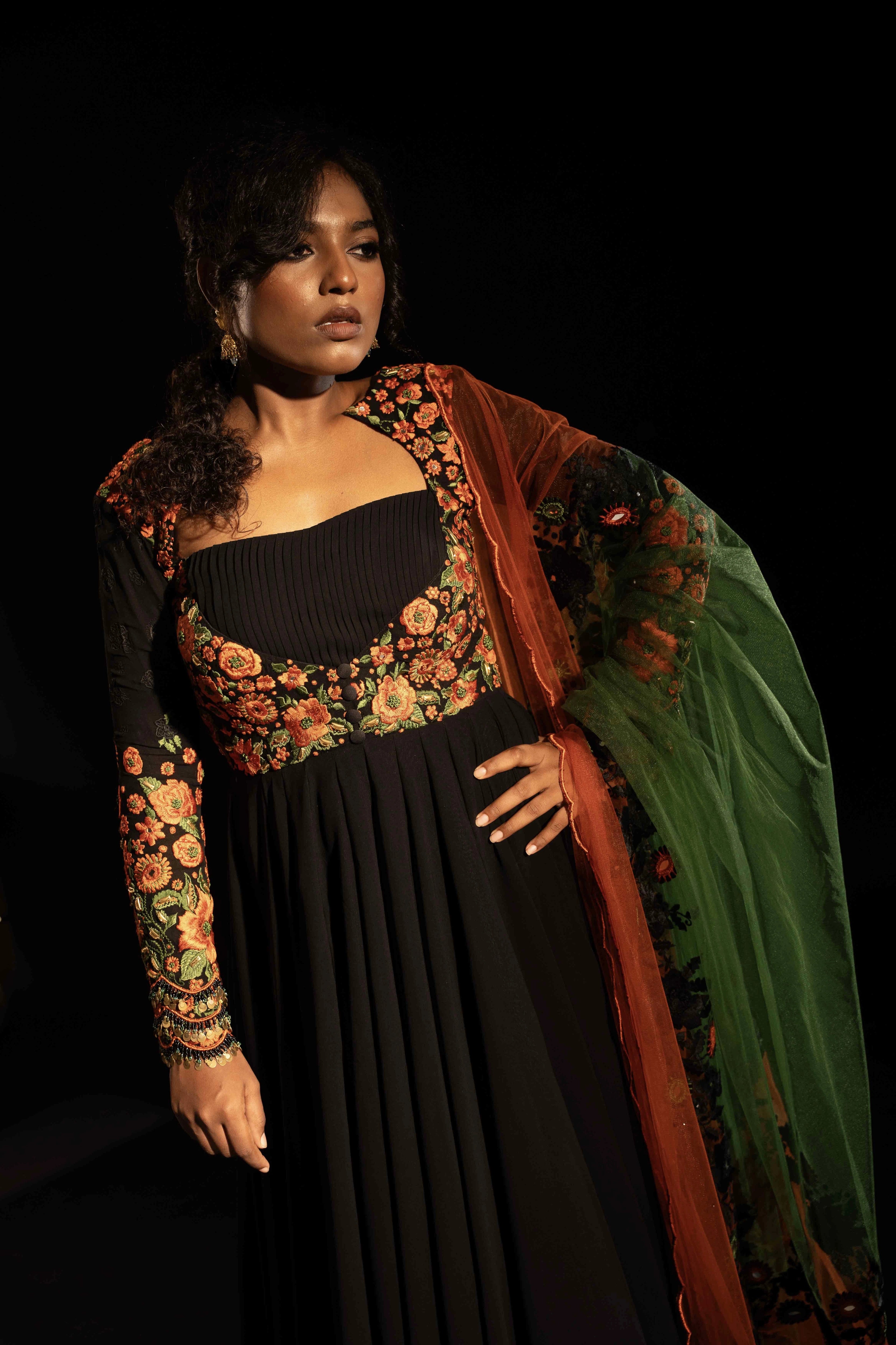 Model Ashika posing for photoshoot in a black anarkali with an orange green dupatta, handcrafted with resham, mirror and metal coins for "HAZE" - A/W'25 capsule collection by Archana Karthick.