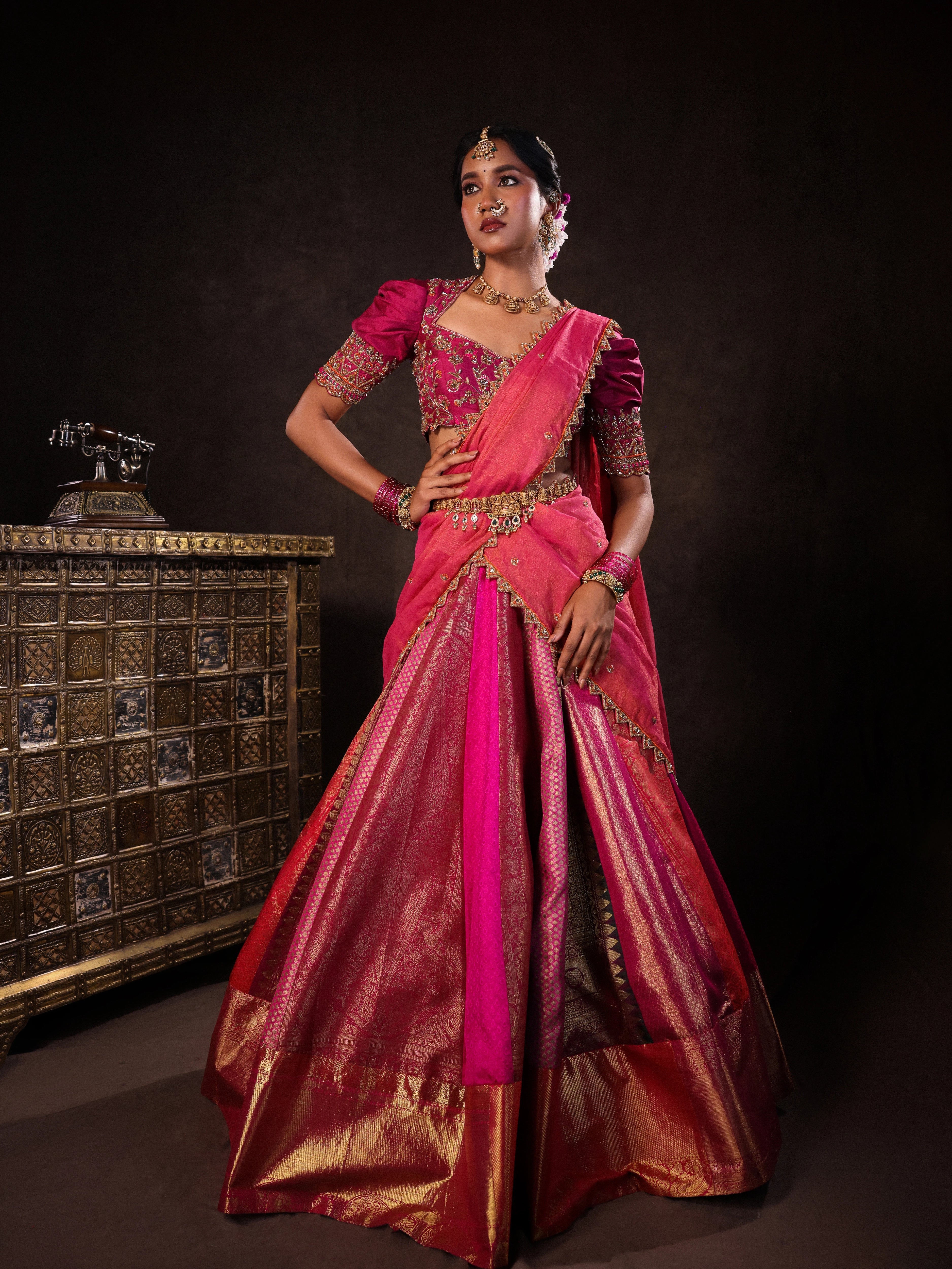 Model Gayathri Dhanasekar poses in a pink & orange silk half saree handcrafted with zardozi, cutdana (bugle beads), resham (silk thread), sequins and rhinestones. Team Archana Karthick completed the style with pink glass bangles, a ring, a choker, jhumkas, nose ring and a maang tikka.