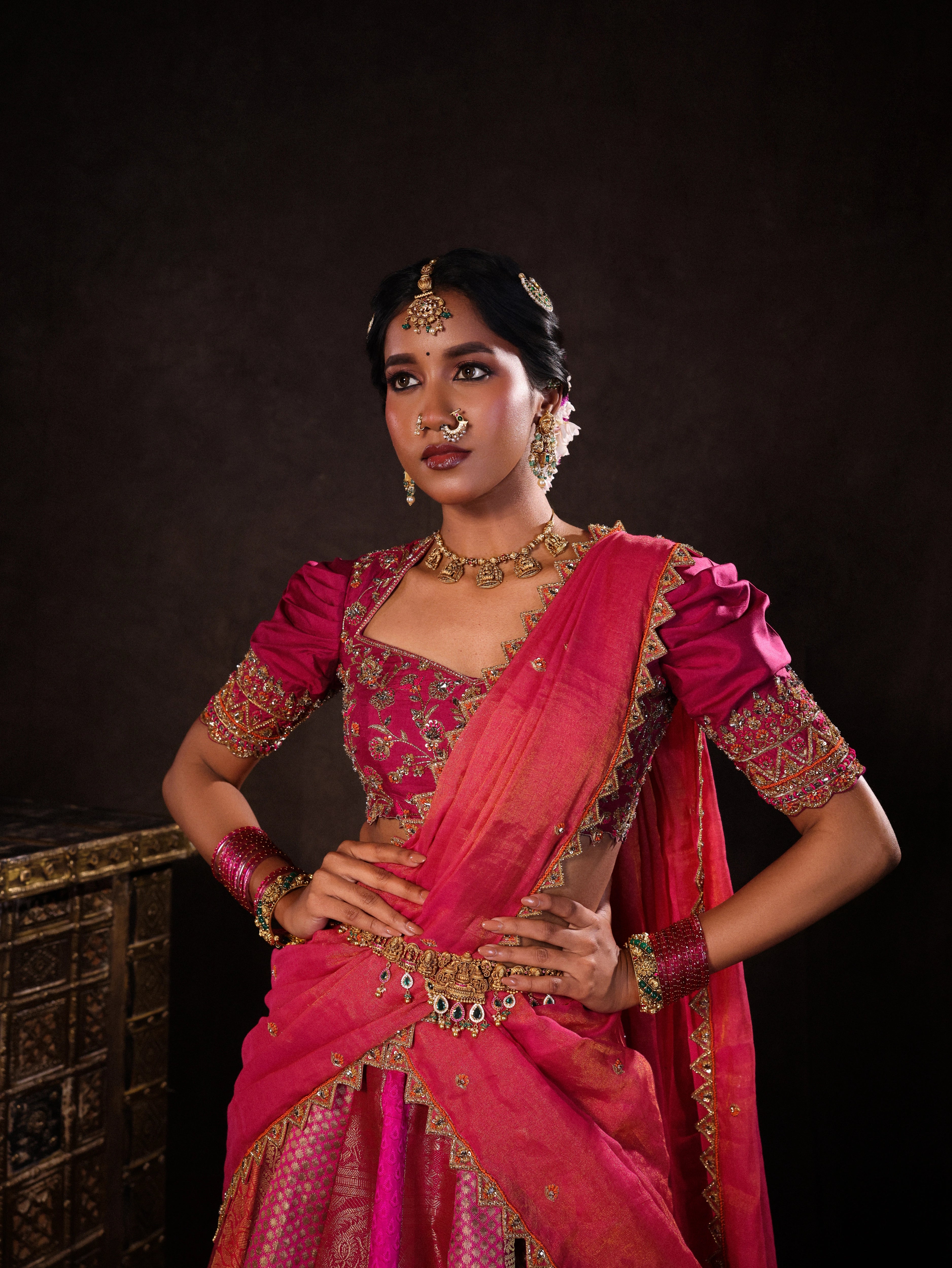 Model Gayathri Dhanasekar posing for front detailing in a pink & orange silk half saree handcrafted with zardozi, cutdana (bugle beads), resham (silk thread), sequins and rhinestones. Team Archana Karthick completed the style with pink glass bangles, a ring, a choker, jhumkas, nose ring and a maang tikka.