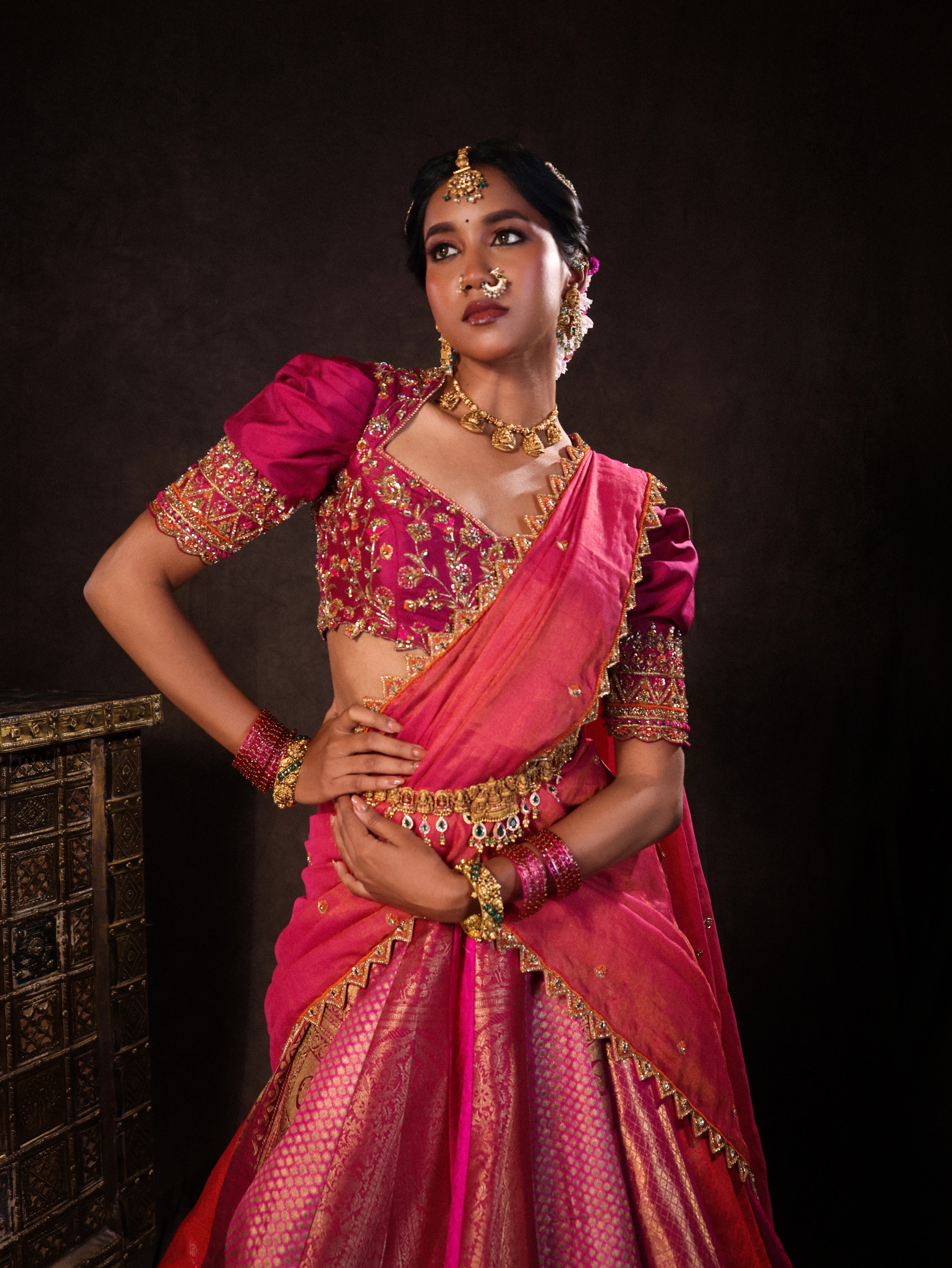 Model Gayathri Dhanasekar posing in a pink & orange silk half saree handcrafted with zardozi, cutdana (bugle beads), resham (silk thread), sequins and rhinestones. Team Archana Karthick completed the style with pink glass bangles, a ring, a choker, jhumkas, nose ring and a maang tikka.